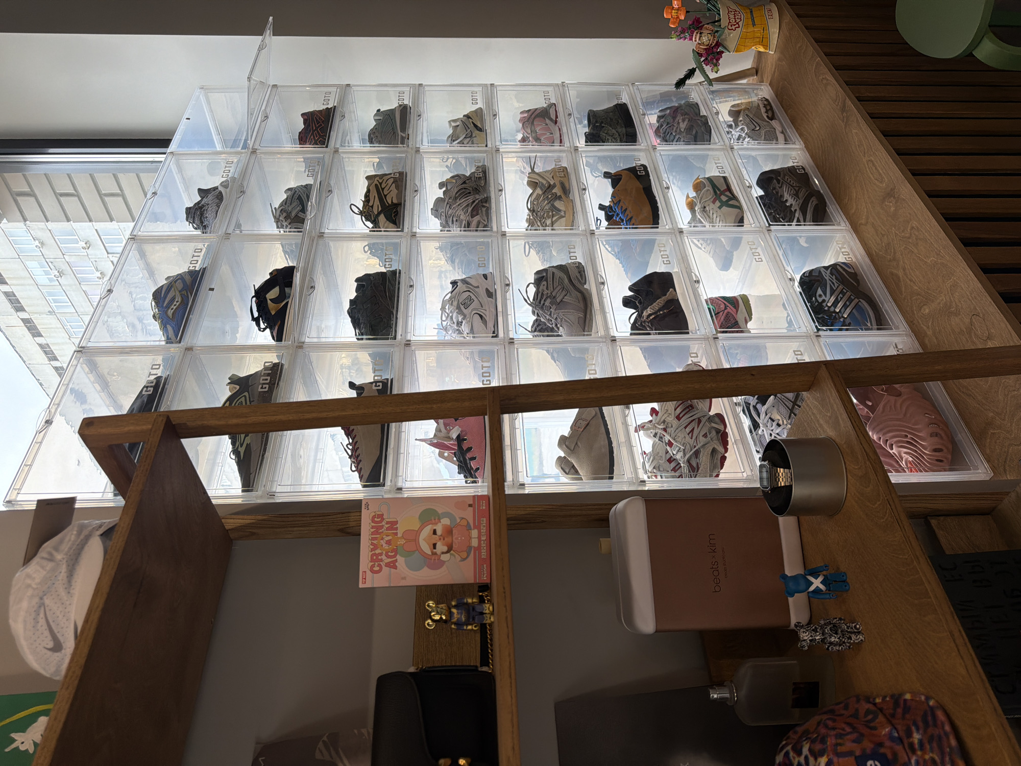 The image displays a collection of sneakers stored in clear plastic boxes arranged in a pyramid shape on a wooden shelf. The sneakers vary in color and style and are neatly organized within the boxes. The boxes are labeled "GOTO." The setting appears to be indoors, with natural light coming from a window through the plastic boxes. The rest of the shelf below the sneakers contains various objects, including a baseball cap, a "Crying Again" book, a "beats x kim" box, a watch, and some small art figures. There is a container of flowers on the top shelf next to the sneakers. The location of the scene is unknown.