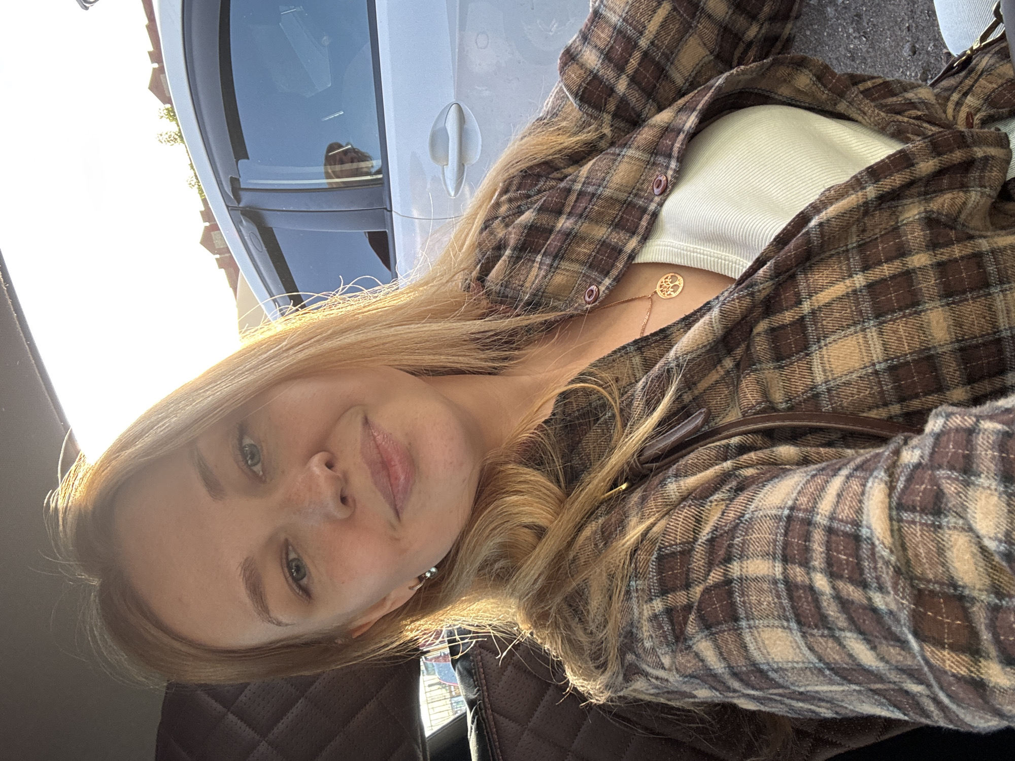 A young woman with long blonde hair and light eyes is captured in a close-up, rotated selfie, likely taken inside a car in Rusanovo, Russia. She wears a brown and beige plaid flannel shirt over a light, possibly cream-colored, ribbed top. A delicate gold chain necklace with a circular "tree of life" pendant adorns her neck. She has a slight, calm smile and appears to have freckles on her nose and cheeks.
Strong, bright sunlight streams from the upper left, creating a luminous halo effect around her hair and strongly illuminating the background. Through the car window, a light-colored vehicle (possibly silver or white) and glimpses of distant buildings and trees are visible, blurred by the intense sunlight. The interior of the car is partially visible, showing dark, possibly quilted leather upholstery on the seats. The time of day appears to be daytime, given the powerful natural light. There is no visible text in the image.