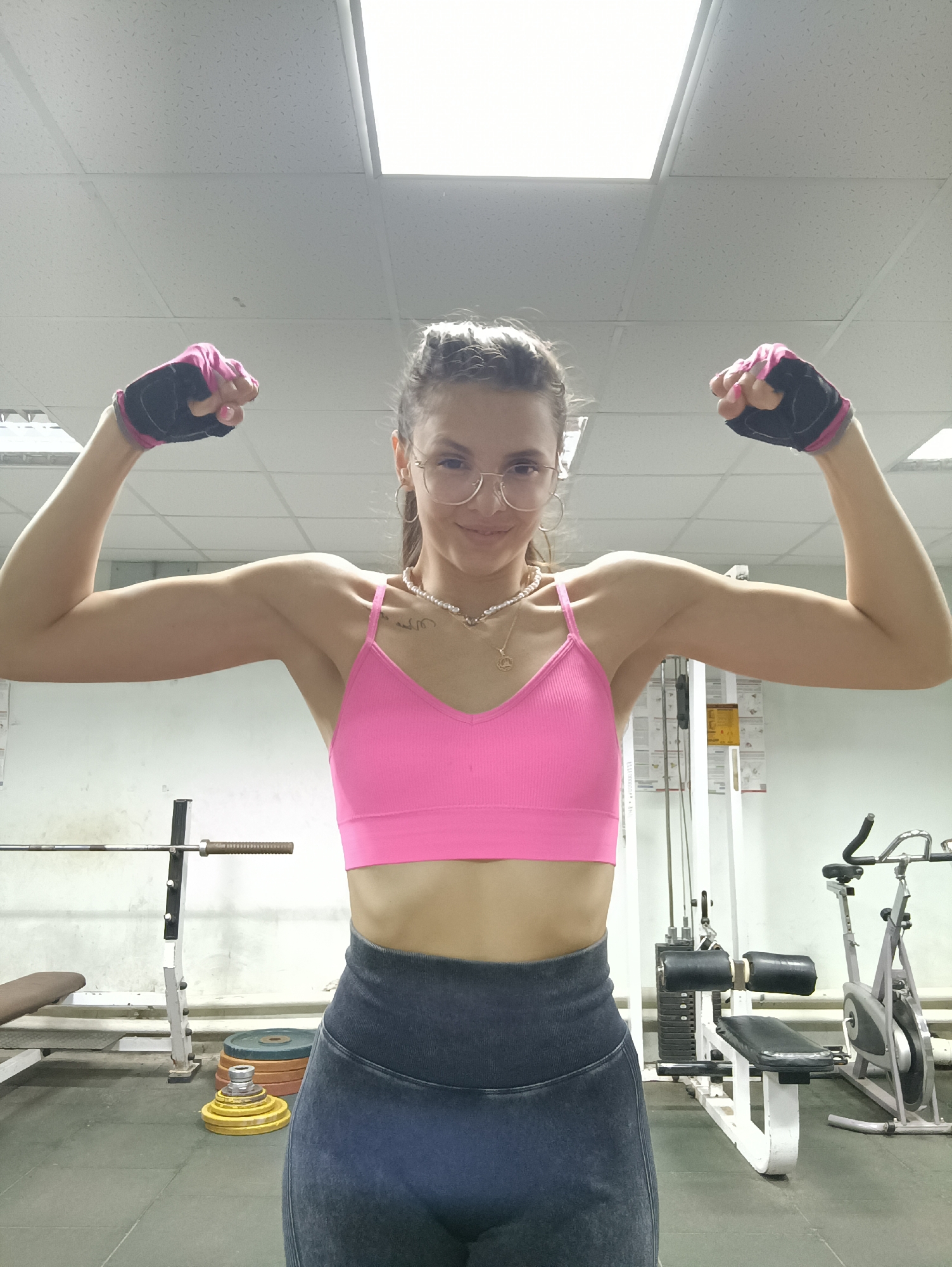 A young woman with her hair pulled back and wearing glasses poses for a selfie in a gym. She is wearing a pink sports bra, dark leggings, and black and pink weightlifting gloves. Her arms are flexed, showing off her biceps. The woman is smiling and looking directly at the camera.
The gym setting includes a weightlifting bench and a rack with weight plates to the left of the woman. To her right, a weight machine and an exercise bike are visible. The lighting suggests it could be daytime or evening, as the overhead fluorescent lights are on. The overall mood is one of strength and confidence. There is no visible text in the image.