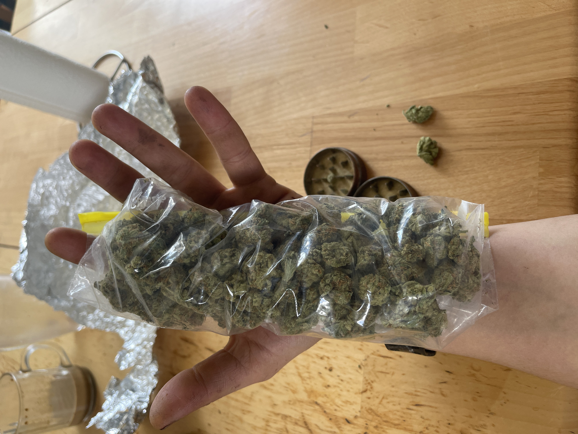 A person's hand holds a clear plastic bag filled with marijuana. The bag is positioned over their forearm. On the wooden surface below, a metal grinder is visible, along with two loose buds of marijuana. In the background, a mug with a brown liquid and crumpled aluminum foil can be seen. The scene appears to be indoors, on a wooden table.