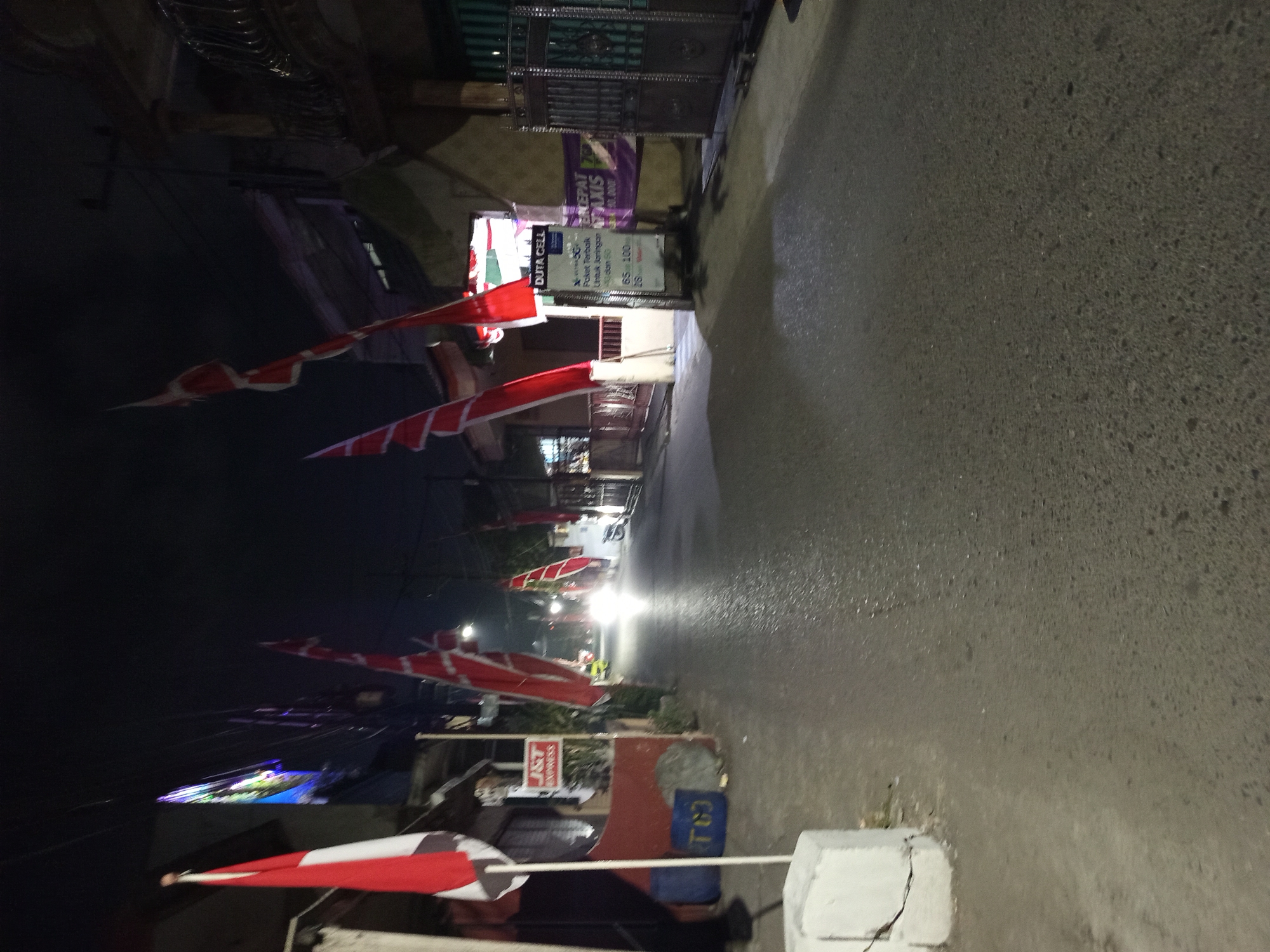 This is a nighttime, slightly blurry, eye-level, wide shot of a street in Tebel, Indonesia. The street is paved with asphalt and appears wet, reflecting the ambient light. Red and white flags are prominently displayed on poles along the street, with one on the left and several further down on the right. A white concrete barrier is visible in the foreground on the left. A sign for "J&T Express" is mounted on a pole on the left side of the street. Further down the street, bright headlights from a vehicle are visible, suggesting traffic. To the right of the street, there are buildings with gates and windows, and a sign that reads "DUTACELL" with promotional text about mobile data packages. The overall atmosphere is that of a residential or commercial street illuminated by artificial light in the evening. No people are clearly visible, but a scooter is parked to the right in the middle ground.