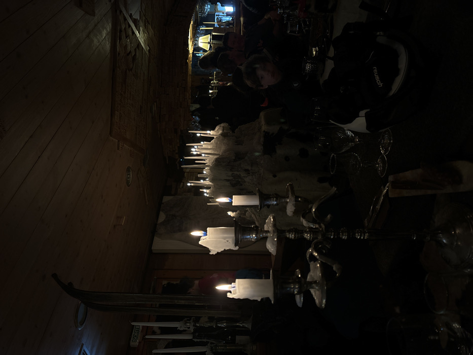 The image captures a dimly lit indoor scene, likely a restaurant or cozy gathering place in the evening or night. The primary light source comes from numerous candles arranged on tables and decorative structures, casting a warm, flickering glow across the room.
In the foreground and mid-ground, multiple candelabras hold pillar and tapered candles, many showing significant melt-down with wax cascading over their sides and holders. One prominent feature is a large, white, textured centerpiece, possibly an ice sculpture or a decorative element resembling snow or rock, studded with many small, upright candles. Wine glasses are visible on the tables, suggesting a dining or social setting.
Several people are seated at tables in the background and mid-ground, mostly silhouetted or partially visible due to the low light. One individual in the mid-ground, wearing a dark jacket, is more discernible, looking towards the right. A "Reusch" logo is visible on an item of clothing or accessory worn by one of the individuals.
The setting appears rustic, with wooden paneling on the walls and what looks like stone or brickwork in the background. The overall ambiance is intimate and atmospheric, created by the abundant candlelight. A sign with the French word "SORTIE" (Exit) is visible on a wooden column or doorframe, confirming a French-speaking location, consistent with Val-d'Isère, France. The scene depicts an event or evening meal, characterized by its warm, candlelit intimacy.