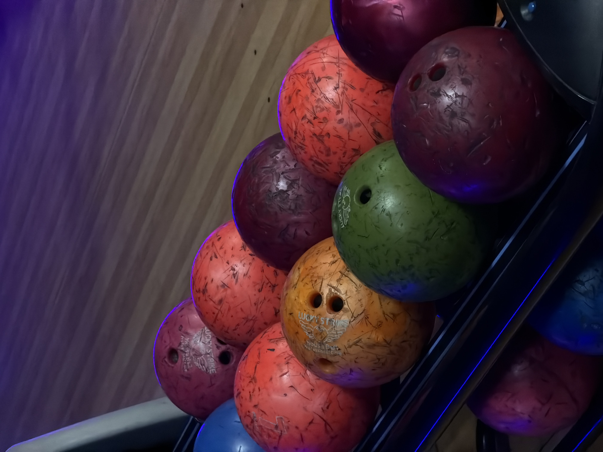This close-up image captures a collection of well-used bowling balls arranged in a dark metal rack, likely within a bowling alley in Izhevsk, Russia. The bowling balls display a variety of colors, including various shades of red, orange, maroon, green, and blue, with several exhibiting visible scuffs and scratches from frequent play.
A distinctive artificial purple and blue light illuminates the left side of the frame, casting reflections onto some of the balls and creating a vibrant, energetic ambiance. The background features vertical wooden panels, possibly part of the bowling lane or an adjacent wall.
Several bowling balls feature visible branding. On a green ball and an orange ball, the text "LUCKY STRIKE" is clearly readable, accompanied by a stylized winged logo. Below this, the words "House Ball" and "Professional Core" are also discernible. Similar, though fainter, text can be seen on a pink/maroon ball in the lower left.
No individuals are present in the image, nor is any specific activity taking place, but the context of the bowling balls in a rack implies an active bowling facility. The artificial lighting suggests an indoor setting, likely during the evening or night.