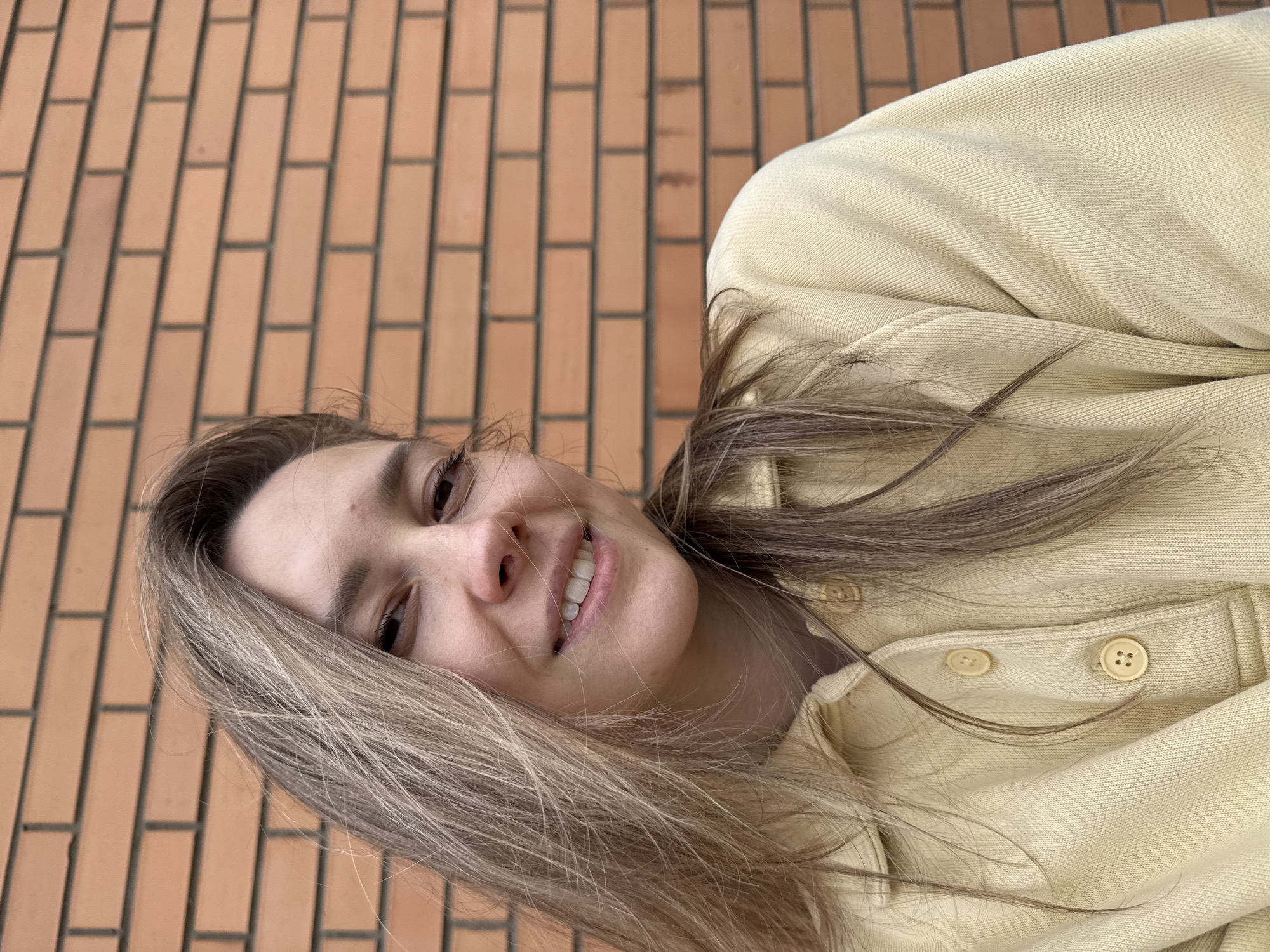 A young woman is smiling at the camera in front of a brick wall. She is wearing a light yellow collared shirt with buttons. The brick wall is made of orange-red bricks laid in a traditional pattern. The photograph appears to be taken in an outdoor setting. The city of Chaikovsky, Russia, cannot be determined from the image.