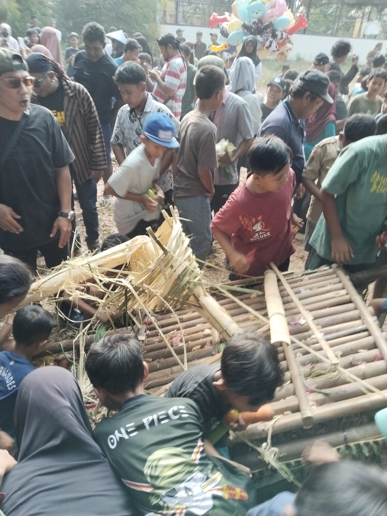 In the village of Karangmalang, Indonesia, a crowd gathers outdoors, with a focus on children. Several children are interacting with a bamboo structure, which appears to be part of a game or activity. One child, wearing a red t-shirt with the word "DELUXE" on it, is positioned near the bamboo structure and is looking downwards. Another child, in a green t-shirt with "ONE PIECE" printed on it, is also visible. Adults are present in the background, some observing the children. The environment suggests a sunny day, with natural lighting and a dirt or sandy ground. The presence of balloons in the background hints at a festive or celebratory occasion.