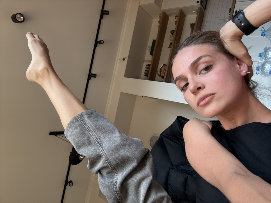An indoor, low-angle shot depicts a young female subject positioned in the lower right portion of the frame, looking directly at the camera. Her head is slightly tilted, with her left hand supporting her jaw; a black smartwatch is visible on her left wrist. Her left leg, partially covered by light grey denim jeans, extends upwards towards the top of the frame, with her bare foot pointed towards the ceiling. The ceiling is white, equipped with a black track lighting system featuring multiple cylindrical spotlights. In the background, a light-colored wall with open wooden shelves displays various small objects, including bottles and a woven basket, above a white cabinet or counter. Several large plastic water bottles are visible on a surface in the lower right foreground, near the subject. A dark, padded garment or jacket is partially visible to the subject's left. The scene is an interior room within the city of Unknown, Unknown.
