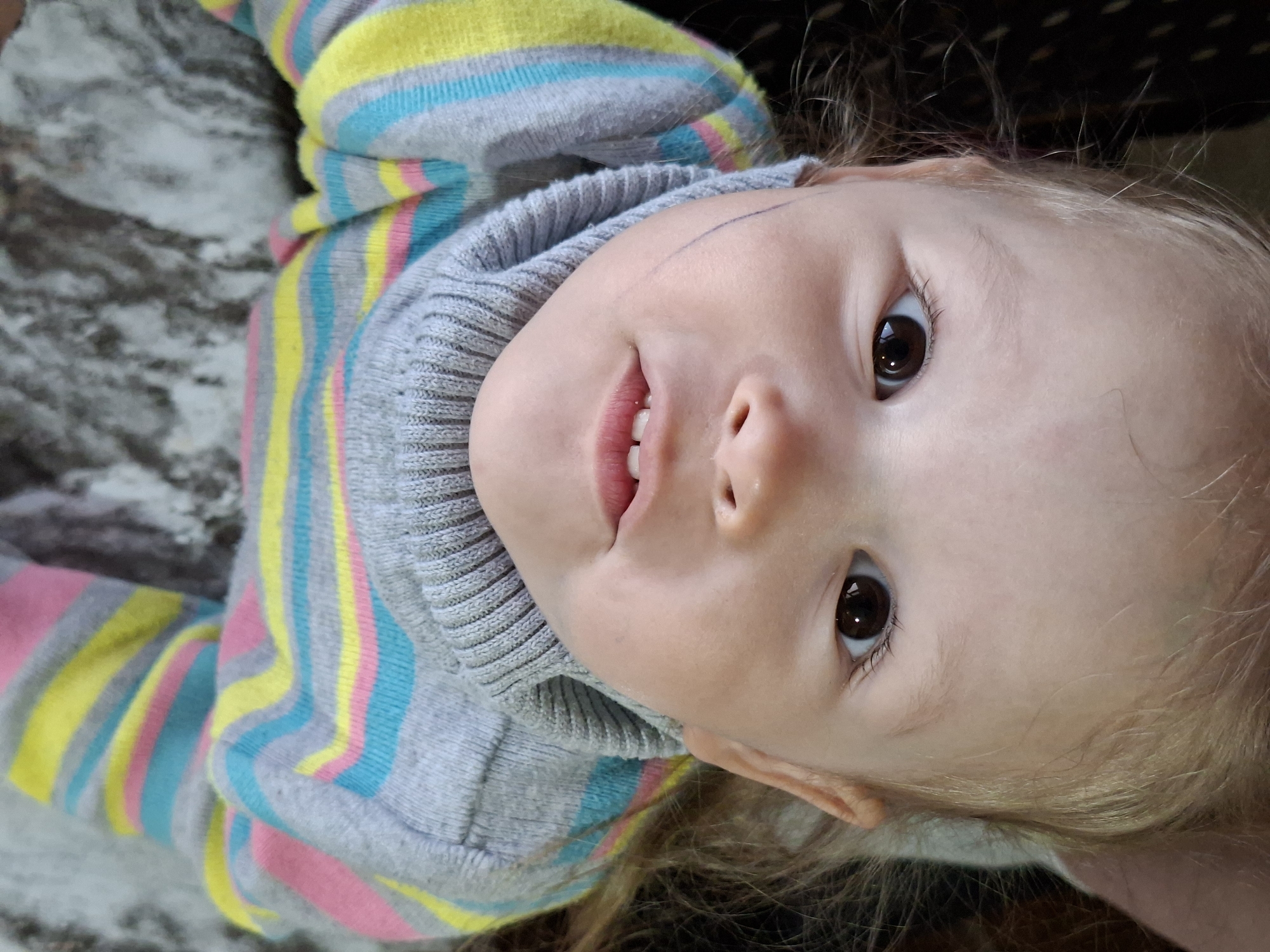 A toddler with fair skin, brown eyes, and light blonde hair is seen from an upward angle, facing the camera. The child is wearing a gray sweater with horizontal stripes of yellow, blue, and pink. A thin, curved line appears on their forehead, possibly a drawing or marking. The background consists of a mottled, light-colored surface to the left and a darker surface to the right. The location is Unknown, Unknown.