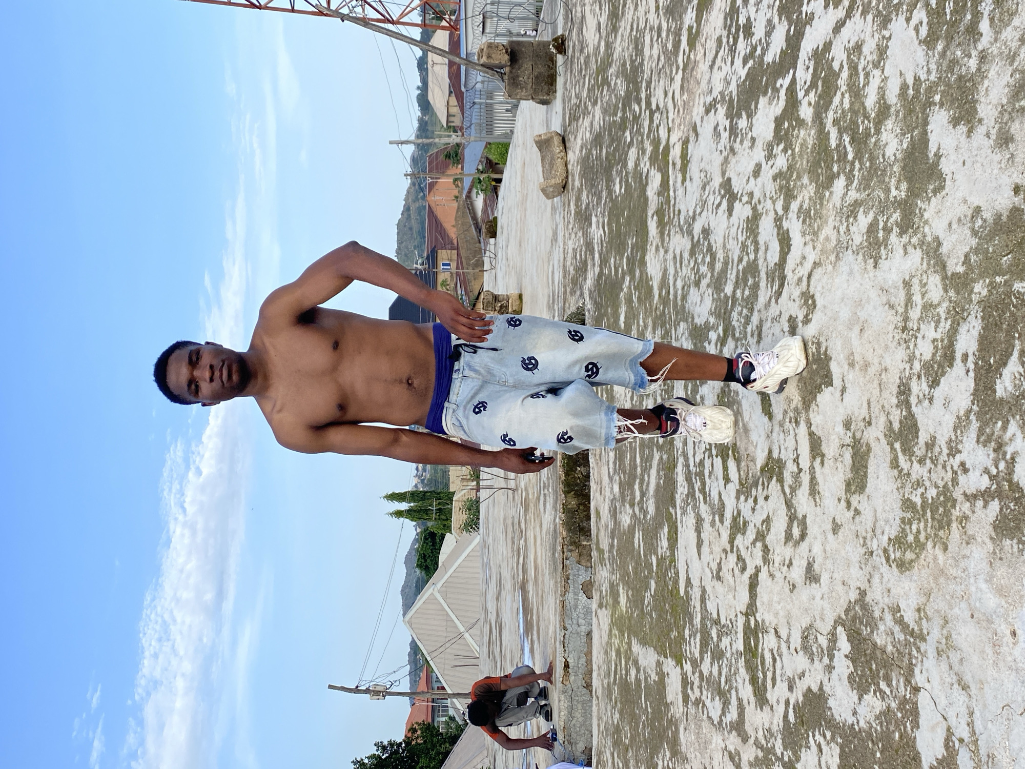 A young Black man stands on a rooftop in Bukuru, Nigeria, facing the camera. He is shirtless, wearing light blue denim shorts with a dark blue waistband and white sneakers. His left leg is slightly raised as if in mid-stride. Behind him, another young Black man is crouched down, appearing to work on the rooftop surface. The rooftop is made of concrete with visible patches of moss or dirt. In the background, there are residential buildings with red roofs, scattered trees, a radio tower, and a hilly landscape under a pale blue sky with scattered white clouds. The overall scene depicts an outdoor urban environment during daylight.