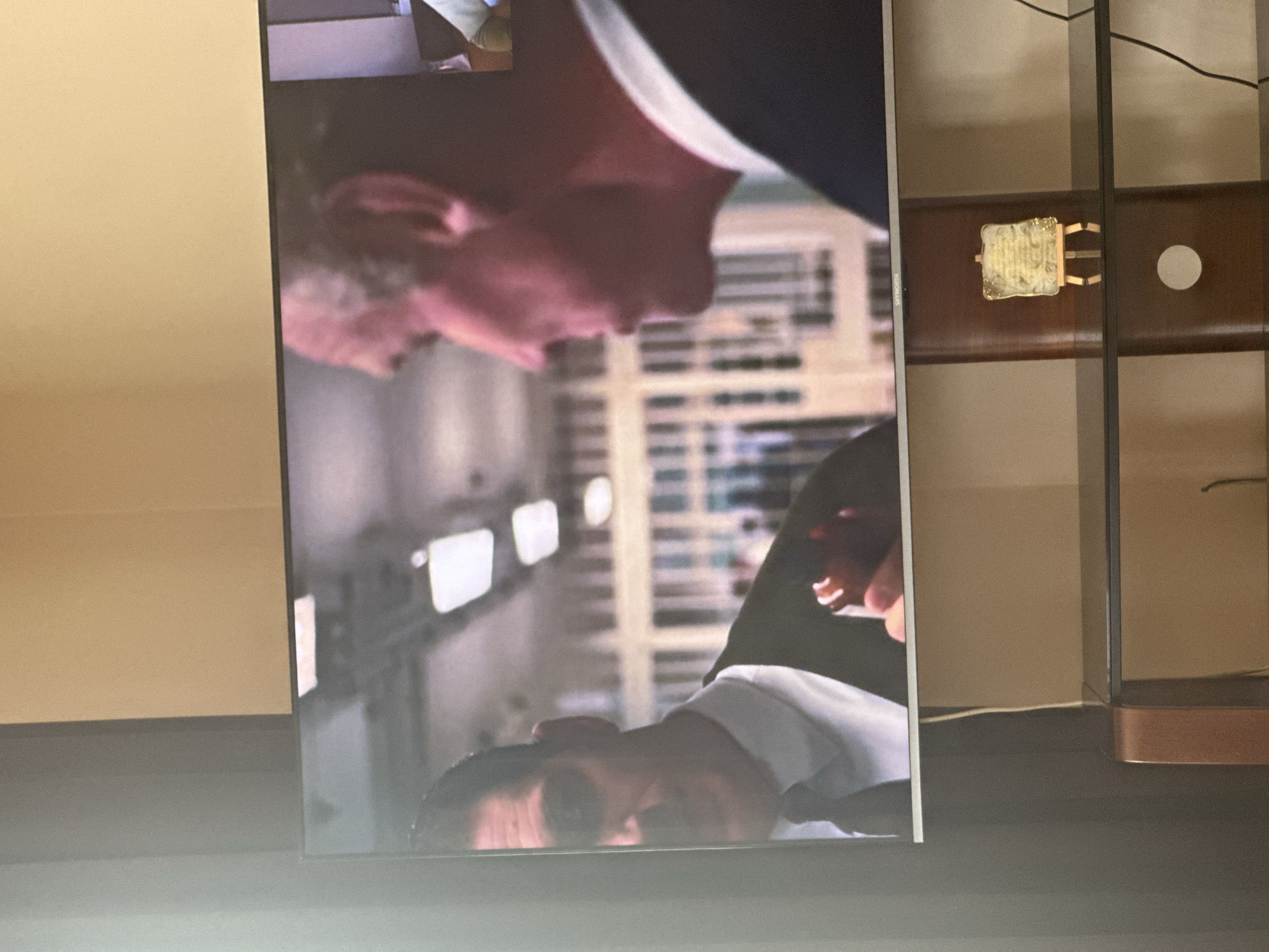 The image shows a television screen displaying a scene with two men in suits, seemingly having a conversation in a bright, possibly office-like environment with window blinds. The screen is positioned against a brown cabinet or shelving unit with a decorative item attached to one shelf. The context of the location in Yerevan, Armenia, cannot be derived from the image.