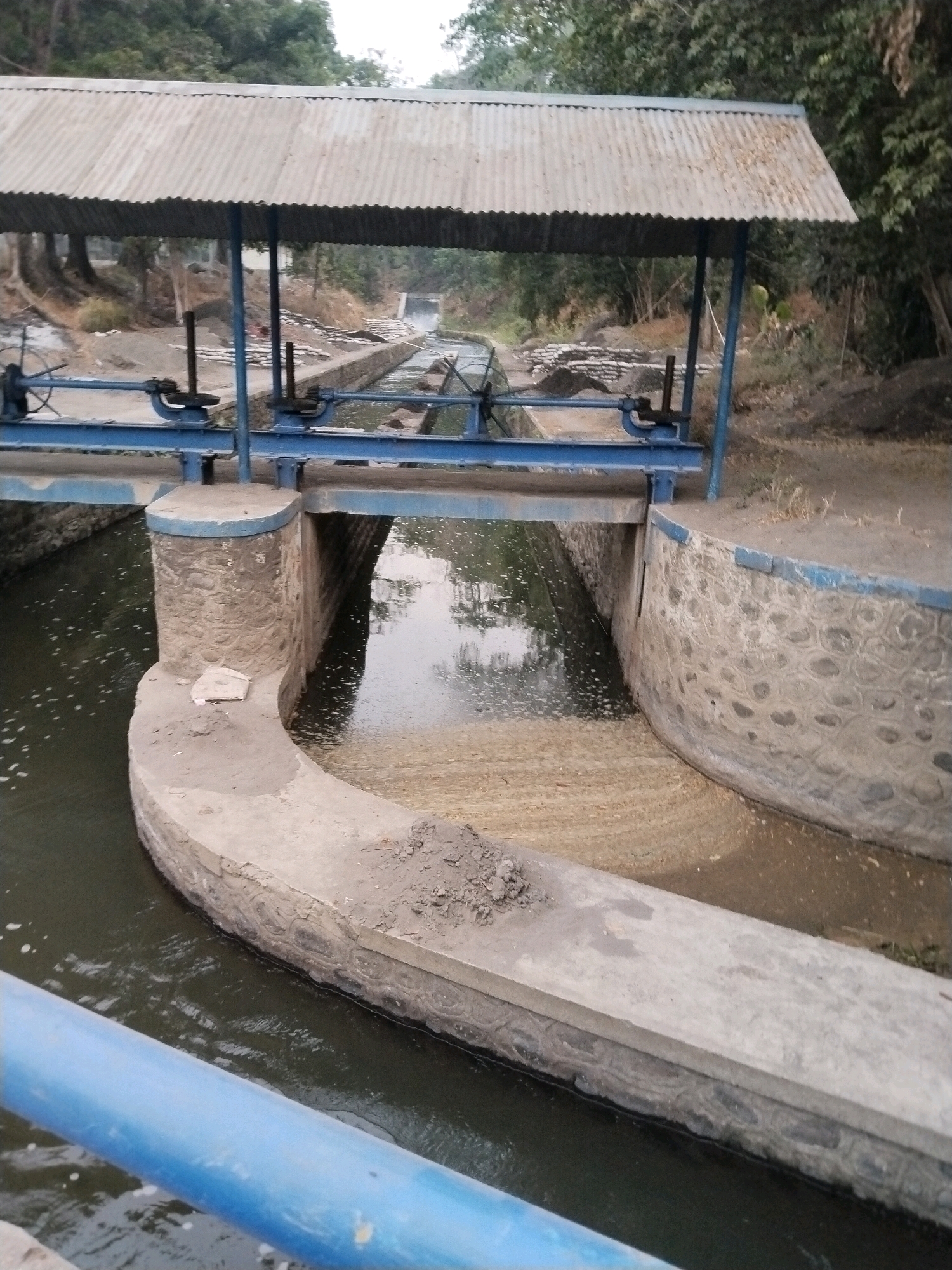 The image shows a canal with concrete walls and a metal roofed structure spanning across it. The structure appears to be a sluice gate mechanism, with blue metal beams and gears visible. The water in the canal is dark and appears to have some debris or foam on its surface. The banks of the canal are lined with concrete and stone. In the background, there are trees and dry vegetation. A blue pipe runs along the bottom left of the image, partially obscuring the view of the canal. The scene is set outdoors, likely in a rural or semi-rural area, given the natural vegetation and the utilitarian nature of the structure. The lighting suggests it is daytime, possibly overcast. The overall impression is of an operational water management system.
