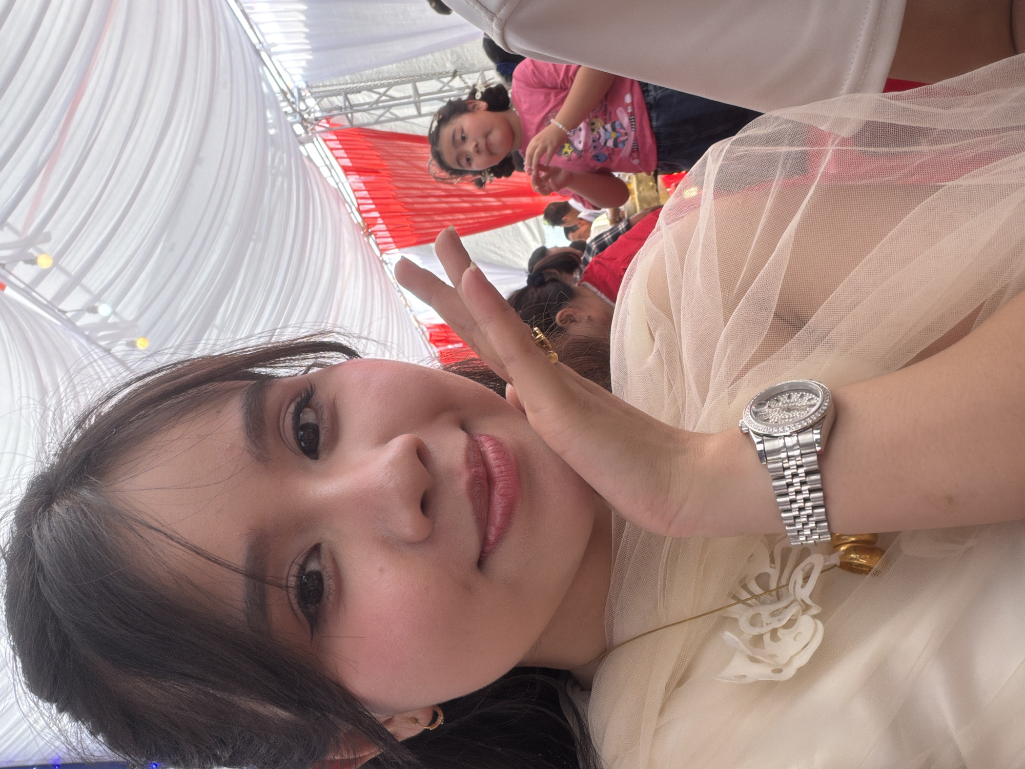 A young woman is captured in a close-up selfie, angled to show her upper body and the surrounding environment, likely in Tay Son Commune, Vietnam. She has dark hair, styled down, and dark eyes, with a subtle smile and pinkish-red lipstick. She wears a light-colored, possibly cream or beige, sheer fabric dress or top, which appears to be layered or ruffled. On her left wrist, she sports a silver-toned wristwatch with a detailed, possibly jeweled, bezel and a metal link band. Her right hand is raised, making a peace sign towards the camera.
In the background, a young girl with dark hair tied in pigtails is visible, looking directly towards the camera with a slight pout or curious expression. She is wearing a pink t-shirt with a colorful graphic print. Other blurred figures of people suggest a gathering or event.
The setting appears to be under a large canopy or tent, with extensive white draped fabric forming the ceiling. Sections of red and white striped fabric are visible in the background, contributing to the festive or event atmosphere. Small, round light fixtures, some yellow and some white, are strung along the canopy structure. The bright, even lighting indicates it is likely daytime. The overall scene suggests a casual event or celebration taking place under a temporary shelter. There is no visible text in the image.