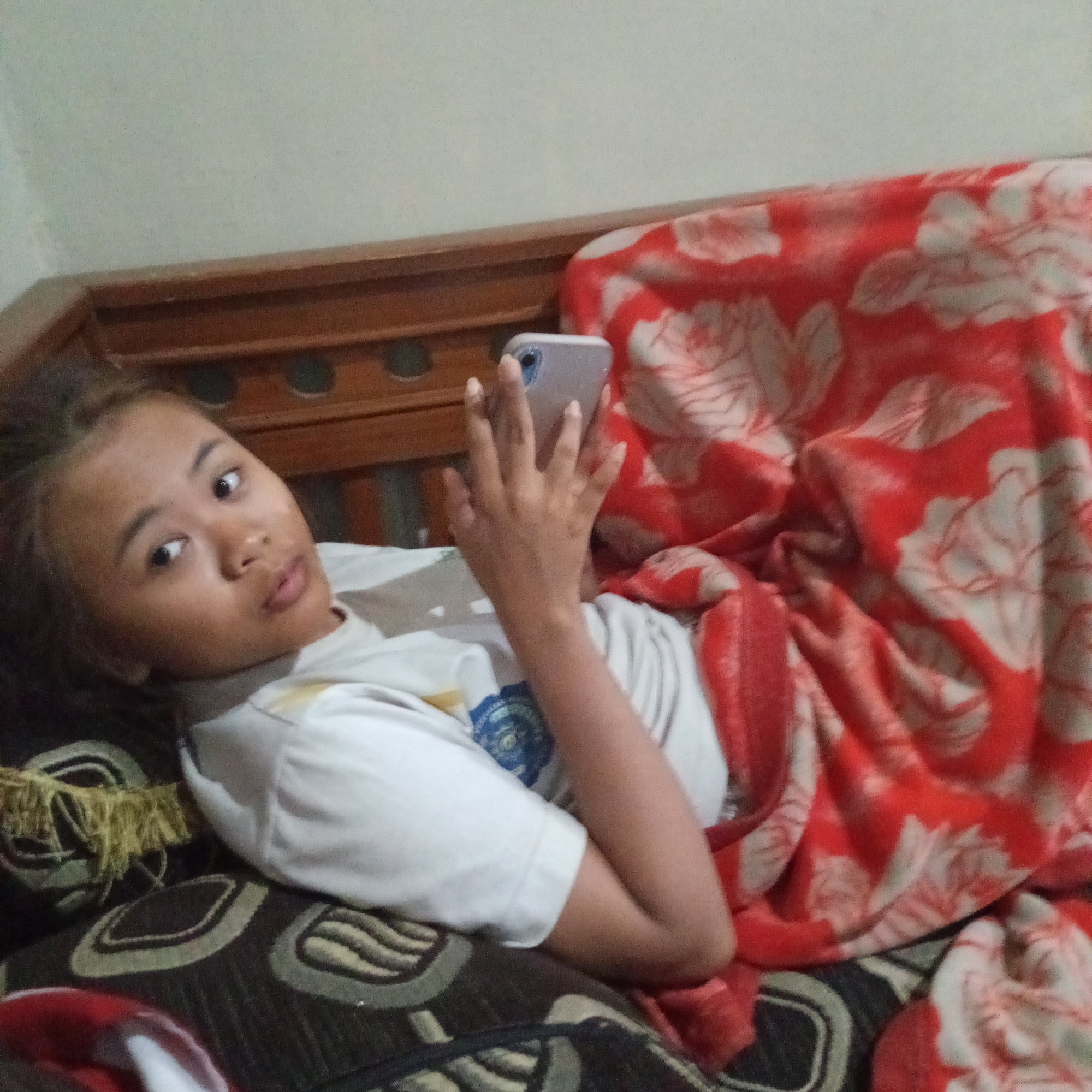 A young female lies on a brown wooden sofa, covered by a red blanket with white floral patterns. She is wearing a white t-shirt and holding a light purple smartphone in her right hand, seemingly looking at it. Her left arm is bent, supporting her head. The background shows a light-colored wall and the wooden structure of the sofa. The image appears to have been taken indoors in a residential setting in Boyolali, Indonesia.