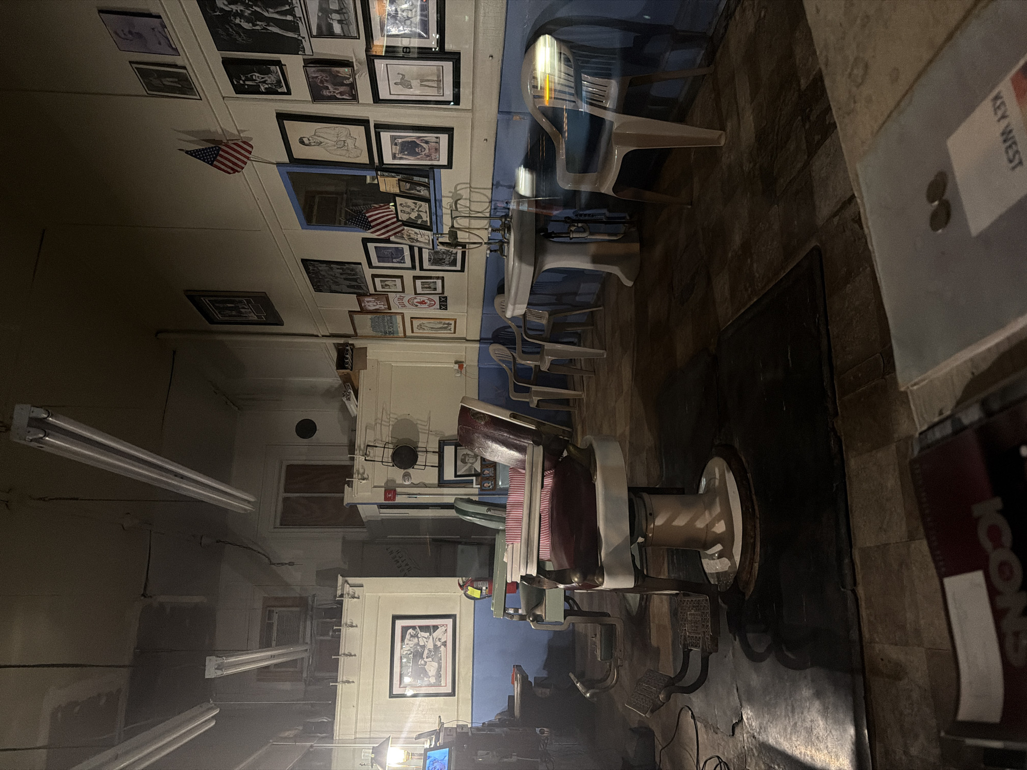 The image shows the interior of a vintage barbershop in Red Bank, United States of America. The scene is dimly lit, with fluorescent lights overhead and a lamp on a side counter providing illumination. The floor is a dark, tiled surface.

Dominating the foreground is a classic barber chair with a red and white striped seat and back, and a white pedestal base. In front of the chair, a black mat is on the floor, and the barber chair's footrest is visible. To the left of the barber chair, a dark wooden counter with a lamp and what appears to be a small screen is situated.

Further into the shop, another barber chair, this one green, is visible. Behind the chairs, a wall is decorated with numerous framed pictures, photographs, and what appear to be sports memorabilia, including an American flag. A sink with a chrome faucet is mounted on the wall, with several plastic chairs arranged around it.

In the background, there is an air conditioning unit mounted high on the wall, and a window covered with boards. The overall atmosphere is one of a well-established, possibly historic, place of business. The image is still and does not capture any ongoing action or interaction.