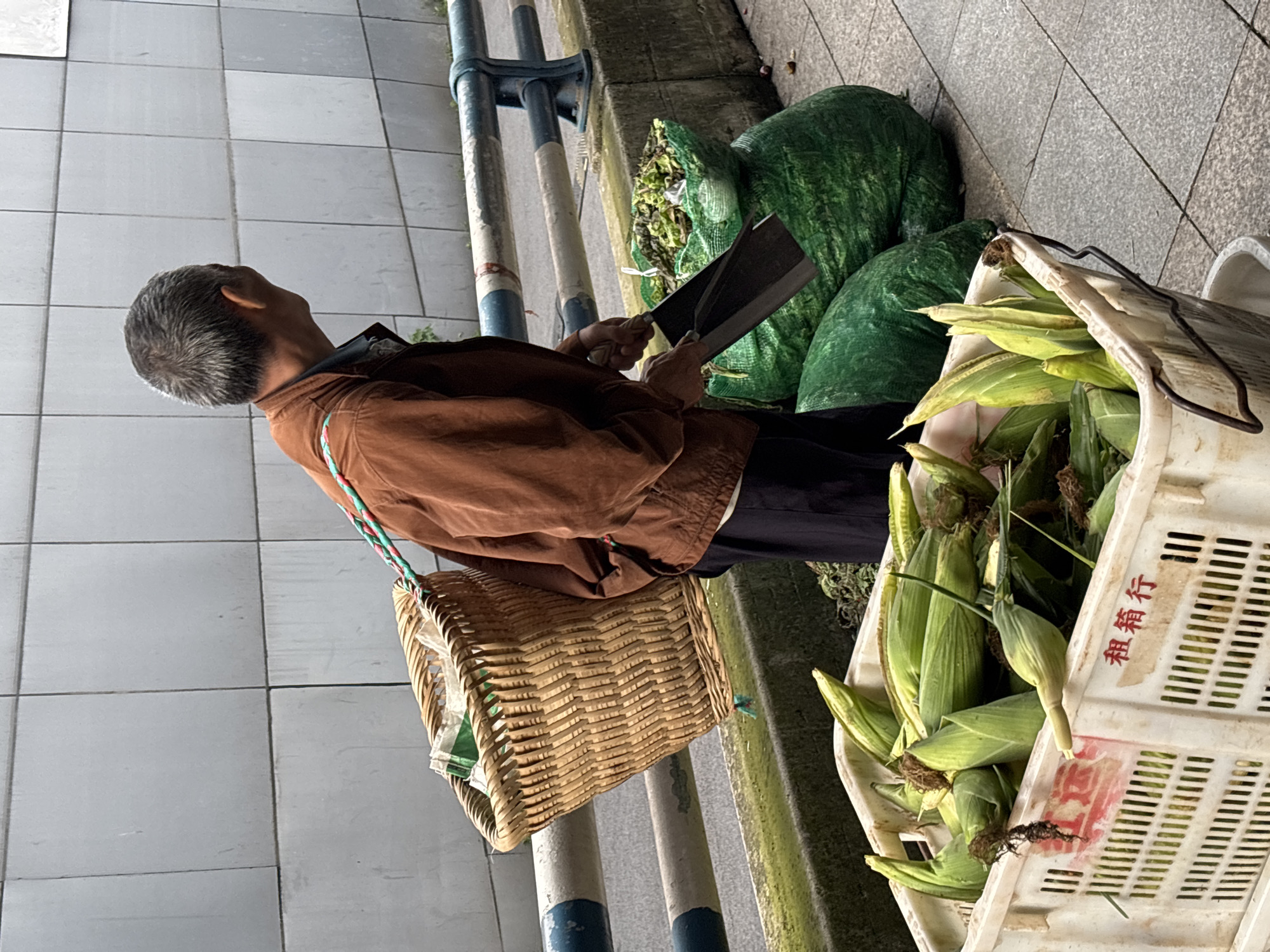 An elderly man with graying hair, wearing a brown jacket and dark pants, stands on a sidewalk in Jiangjin, China. He has a woven basket slung over his shoulder, containing what appears to be paper or cloth. In front of him, he is using a dark-colored shovel to scoop green leafy material from a large green bag into another green bag. Next to the bags, a white plastic crate is filled with fresh corn on the cob. The background consists of a gray tiled wall and a metal railing.