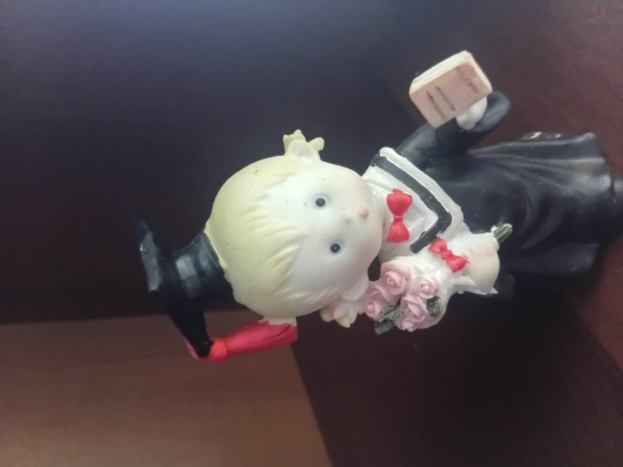 A small, ceramic figurine of a child dressed in graduation attire stands on a dark, wooden surface. The child has blonde hair styled into two pigtails, with a black graduation cap and a red tassel. They are wearing a black graduation gown with a white collar and a red bowtie. In their left hand, the figurine holds a bouquet of pink roses, and in their right hand, a small, open book with text. The background is dark and out of focus, suggesting an indoor environment, potentially a shelf or tabletop in a home. The location is not determinable from the image.