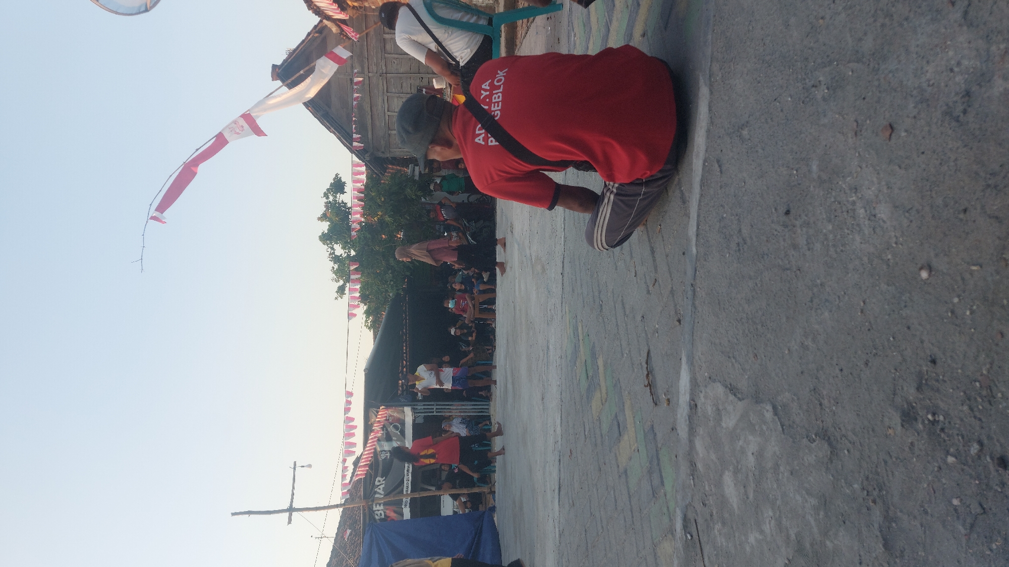 In Jatirogo, Indonesia, a man wearing a red shirt and a grey cap sits on the ground, facing away from the camera, observing an outdoor gathering. The gathering appears to be taking place on a concrete ground, possibly a street or a court, with a building and trees in the background. Red and white Indonesian flags are prominently displayed, strung across the area and attached to poles, indicating a celebration or national event. People are visible in the background, some standing, others seated, suggesting a community event. The lighting indicates it is daytime, likely late afternoon or early morning given the soft sunlight.