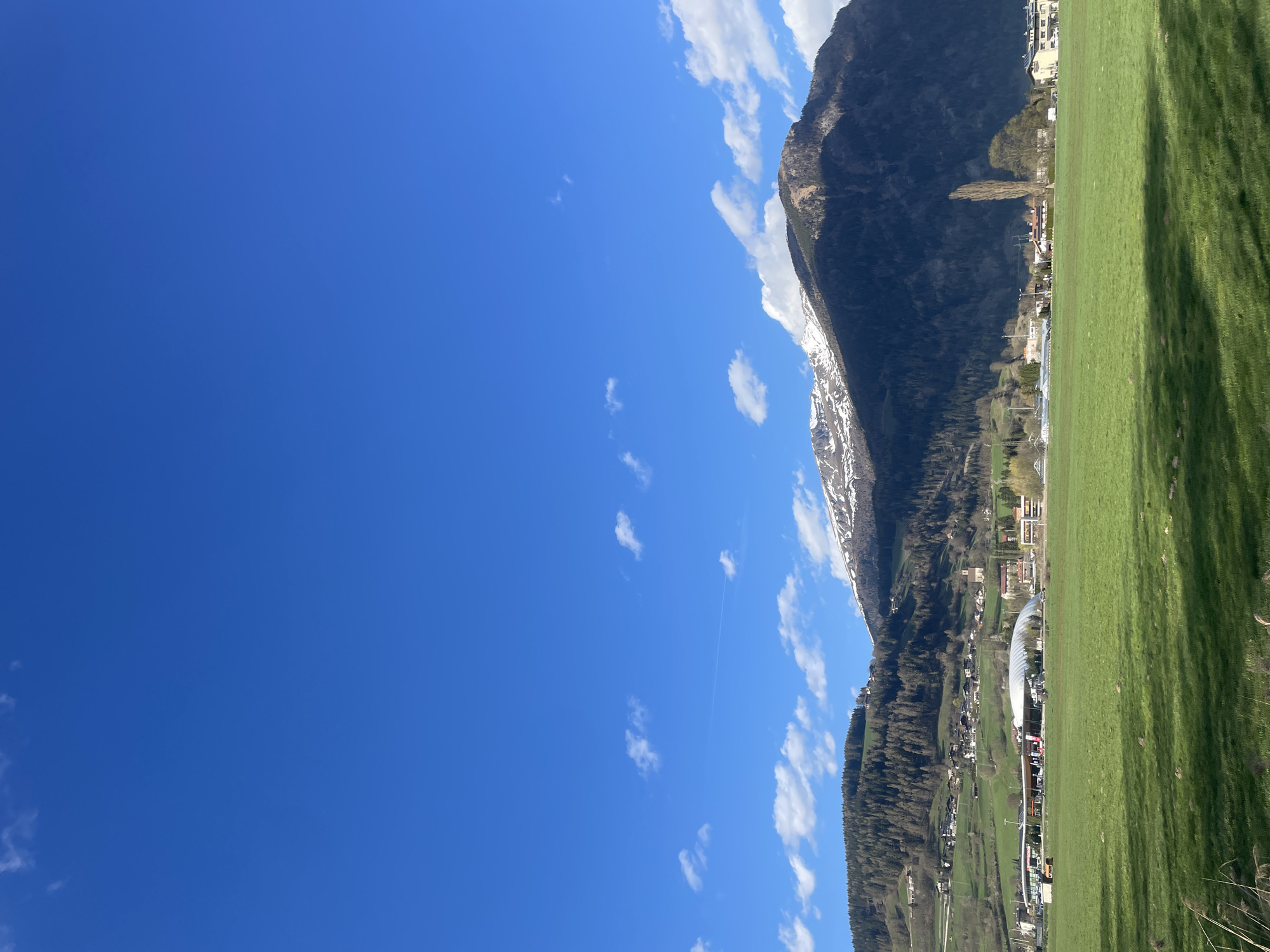 The image depicts a view in Sterzing-Vipiteno, Italy. The foreground consists of a bright green grass field. Behind the field, there's a town with buildings, including a prominent arched structure. A mountain range rises behind the town, partly covered with patches of snow. The sky is a clear blue, dotted with scattered clouds. There appears to be a contrail in the sky.