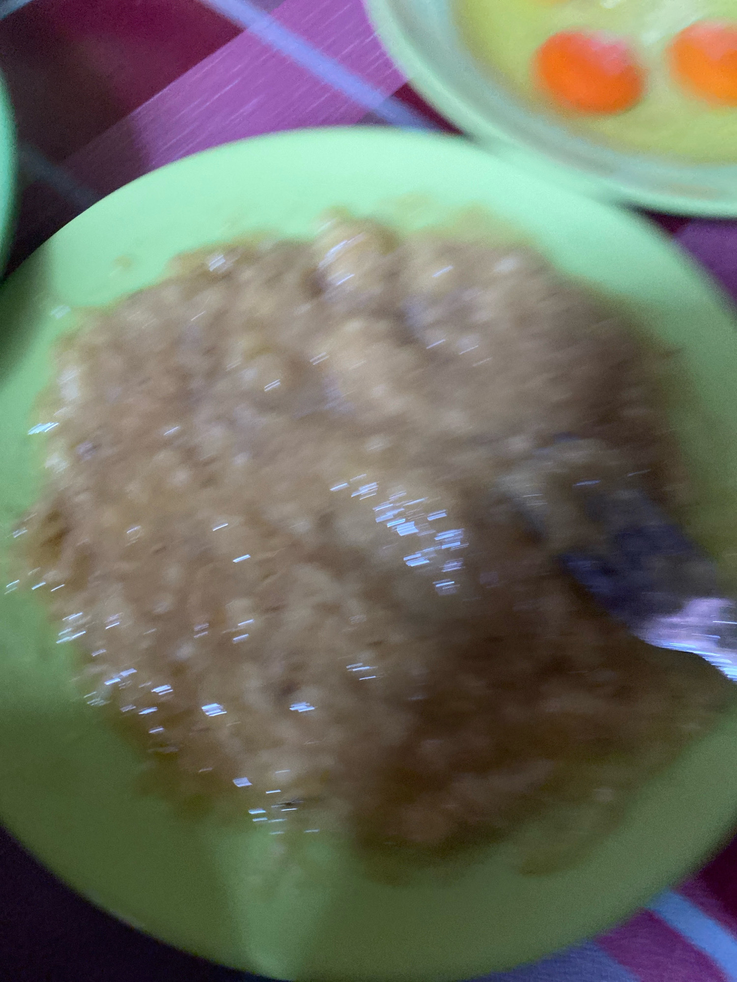 The image shows a close-up of a light green plate containing mashed food, possibly fruit or vegetables, with small white highlights scattered across its surface, suggesting a slight shine or reflection. A dark, possibly metallic object, such as a spoon or fork, is partially submerged in the mashed food on the right side of the plate. The plate is positioned on a surface covered with a fabric that has a checkered pattern of red and purple stripes. In the background, another light green plate is visible, containing what appears to be egg yolks in a yellowish liquid. The overall scene suggests a meal or food preparation in progress. The image quality is blurry, indicating motion during capture. There is no discernible information within the image that would allow for the identification of a specific location in Banjarmasin, Indonesia.