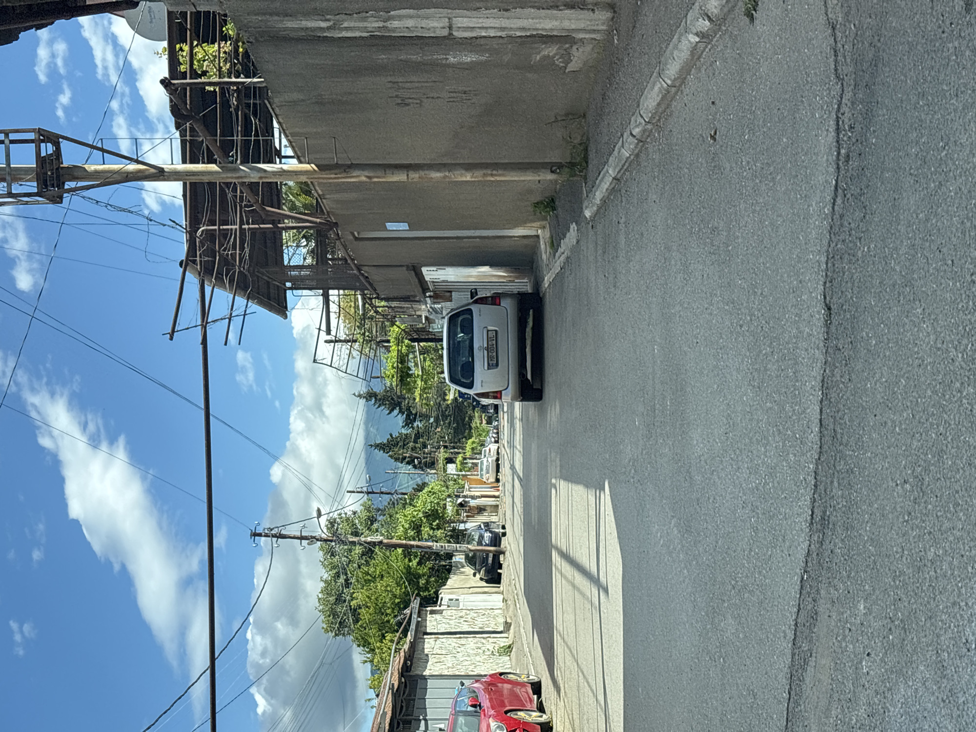 The image captures a narrow street in Tbilisi, Georgia, characterized by a mix of residential and utility features. The street is paved and exhibits wear. It is flanked by buildings on both sides; one side has a series of connected, aged concrete structures, possibly garages, with a white van parked beside one of them. The opposite side shows houses and fences, including a red car parked near the camera. Overhead, utility poles with power lines stretch across the sky, contrasting with the backdrop of trees and distant mountains. The sky is partly cloudy, suggesting daylight conditions.