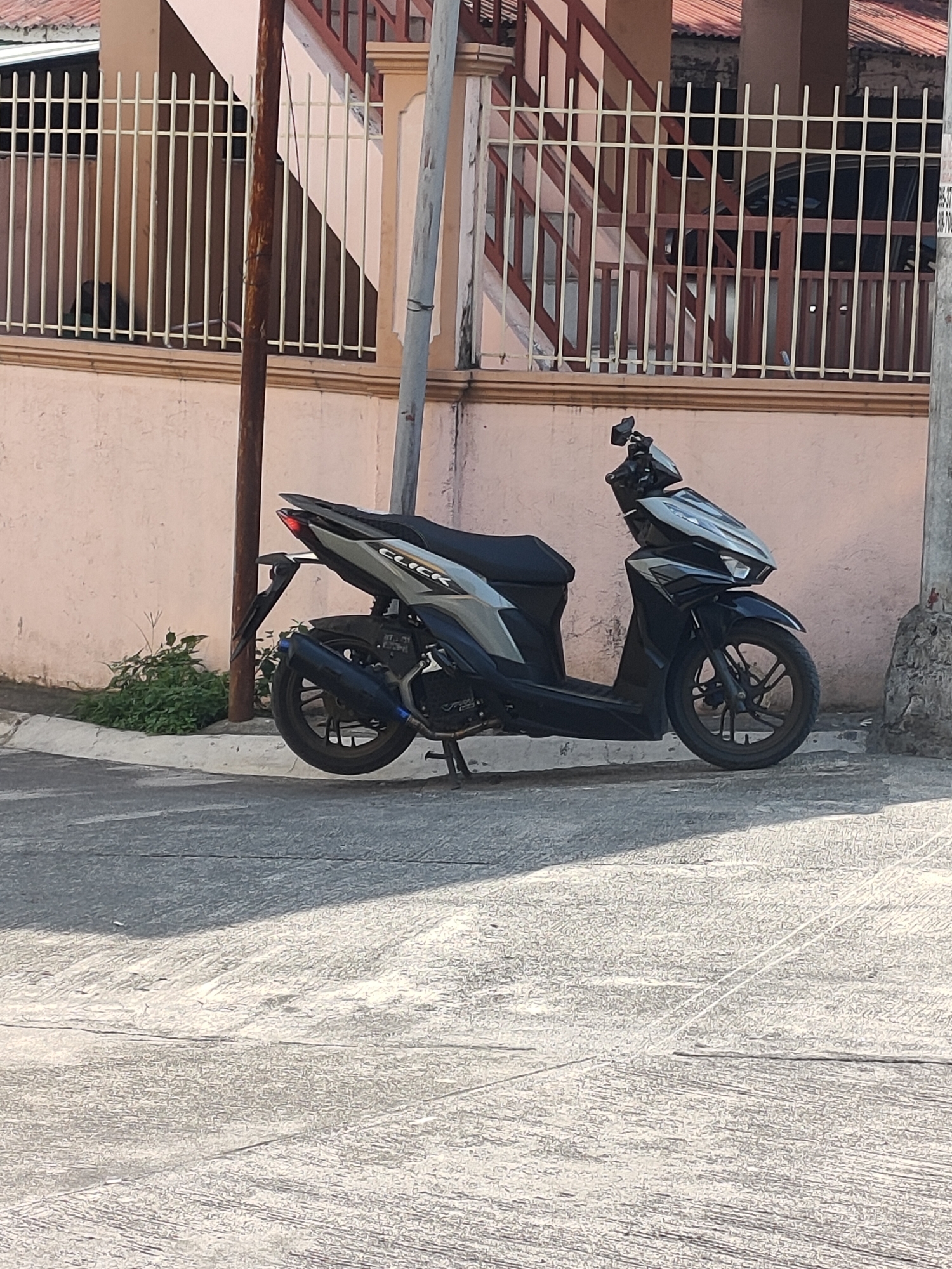 A black and silver Honda Click scooter is parked on a concrete road next to a beige wall and a white metal fence. The scooter is angled to the right, with its rear tire closest to the viewer. The scooter has a dark blue exhaust pipe with a chrome accent. Behind the scooter, there is a patch of green bushes. A utility pole stands to the right of the scooter. In the background, a building with a reddish-brown roof and a brown and white railing is visible. The scene appears to be in a residential area in San Pascual, Philippines, as indicated by the scooter model and the architectural style.