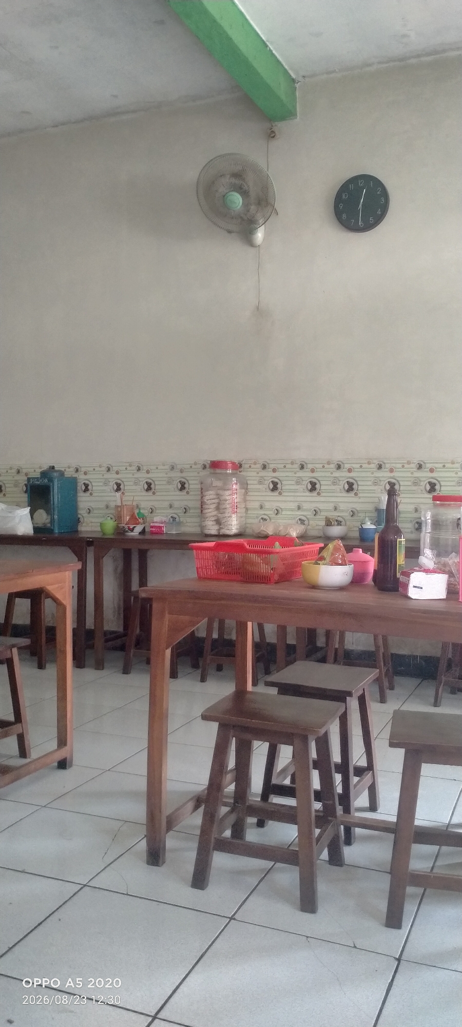 This is a photograph of a simple dining area, likely within a restaurant or food stall, located in Dusun Satu, Indonesia. The scene is dominated by wooden tables and stools, with a tiled floor and a light-colored wall. On the wall, a ceiling fan is visible in the upper left quadrant, and a black circular clock with white hands and numbers is to its right. The clock indicates a time between 12:30 and 12:35. A decorative border with a repetitive pattern runs horizontally across the upper portion of the wall, above a darker horizontal band. On the tables, there are several items including a blue electrical appliance, jars of food, a red plastic basket containing what appears to be snacks or food items, and a brown glass bottle. The lighting suggests it is daytime. A timestamp in the lower left corner reads "OPPO A5 2020 2026/08/23 12:30". There are no people visible in the image.