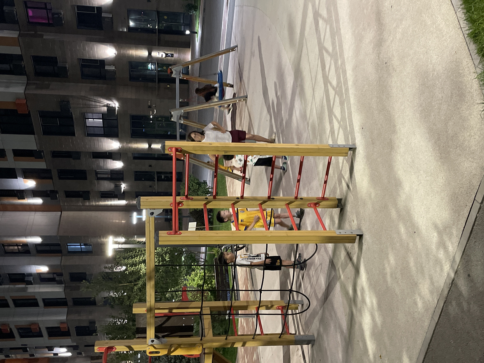 A night-time exterior shot shows a modern playground in front of a multi-story residential building. The building, with its contemporary facade and abundant outdoor lighting, is located in Almaty, Kazakhstan. In the foreground, a climbing structure made of yellow wood and red metal bars is the central focus. Three young boys are near the structure. One boy, wearing a white t-shirt with graphics and black shorts, stands in the center of the climbing frame. To his right, another boy in a yellow soccer jersey and shorts walks away from the climbing structure. A third boy in a white t-shirt and patterned shorts is partially visible on the far right, near the climbing structure. In the middle ground, a woman in a striped t-shirt and burgundy shorts stands with her hands on her hips, looking towards the playground. Behind her, a swing set is visible with a child on a blue swing. The playground area is paved with light-colored concrete, and shadows from the playground equipment are cast across the ground. Lush green trees are present behind the climbing structure, and the surrounding area is illuminated by artificial lights from the building and street lamps.