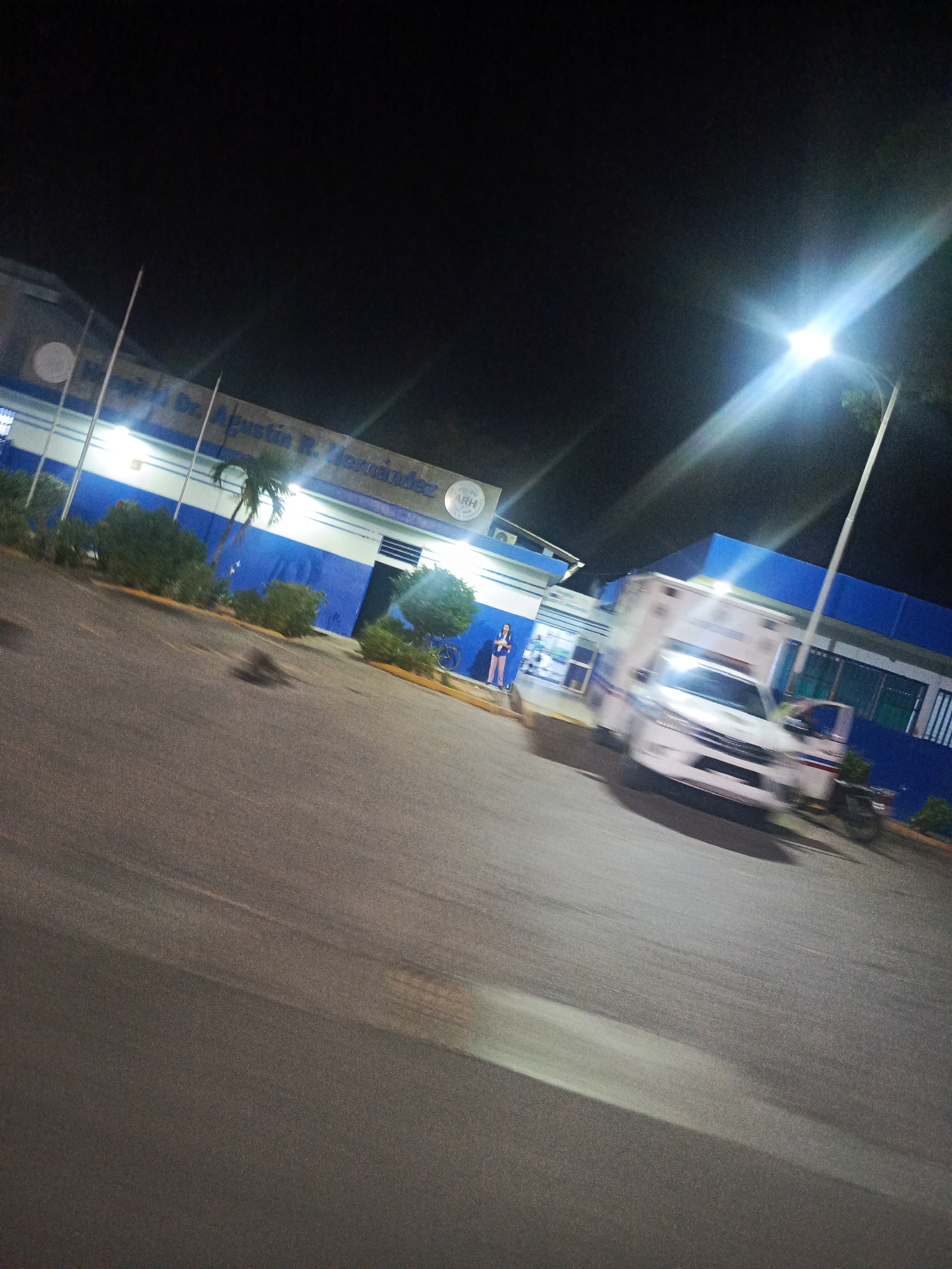 The image shows the exterior of a building at night. The building is lit by artificial lights and appears to be a hospital, with signage that reads "Hospital Dr. Agustín R. Hernández". In front of the building, there is a white ambulance, which appears to be in motion due to motion blur. A person in a blue top and pink pants is standing near the entrance of the building. There are also several trees and bushes in front of the building. The ground in front of the building is paved and appears to be a parking lot. A streetlight illuminates the scene from the right side of the image. The image is taken from a slightly elevated angle and is blurred, suggesting it was taken from a moving vehicle or with a shaky hand. The location is identified as the city of Juan Griego, Venezuela.