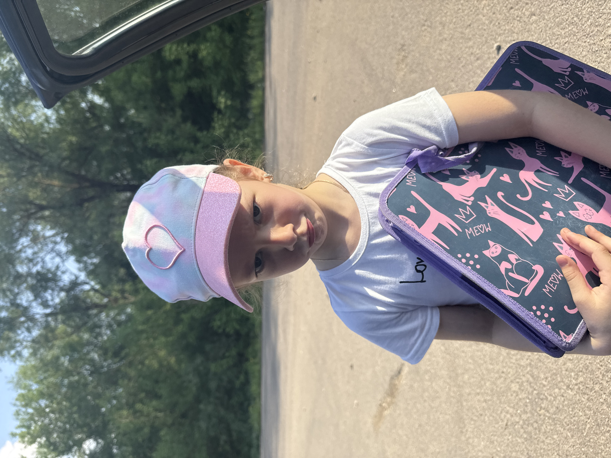 A young girl stands outdoors, holding a folder with a cat pattern. She wears a white T-shirt and a hat with a heart. Part of a car is visible in the upper left corner. The setting is outdoors with trees and a road in the background. It is difficult to definitively confirm the location as Shevlyakovo, Russia based on the image alone.