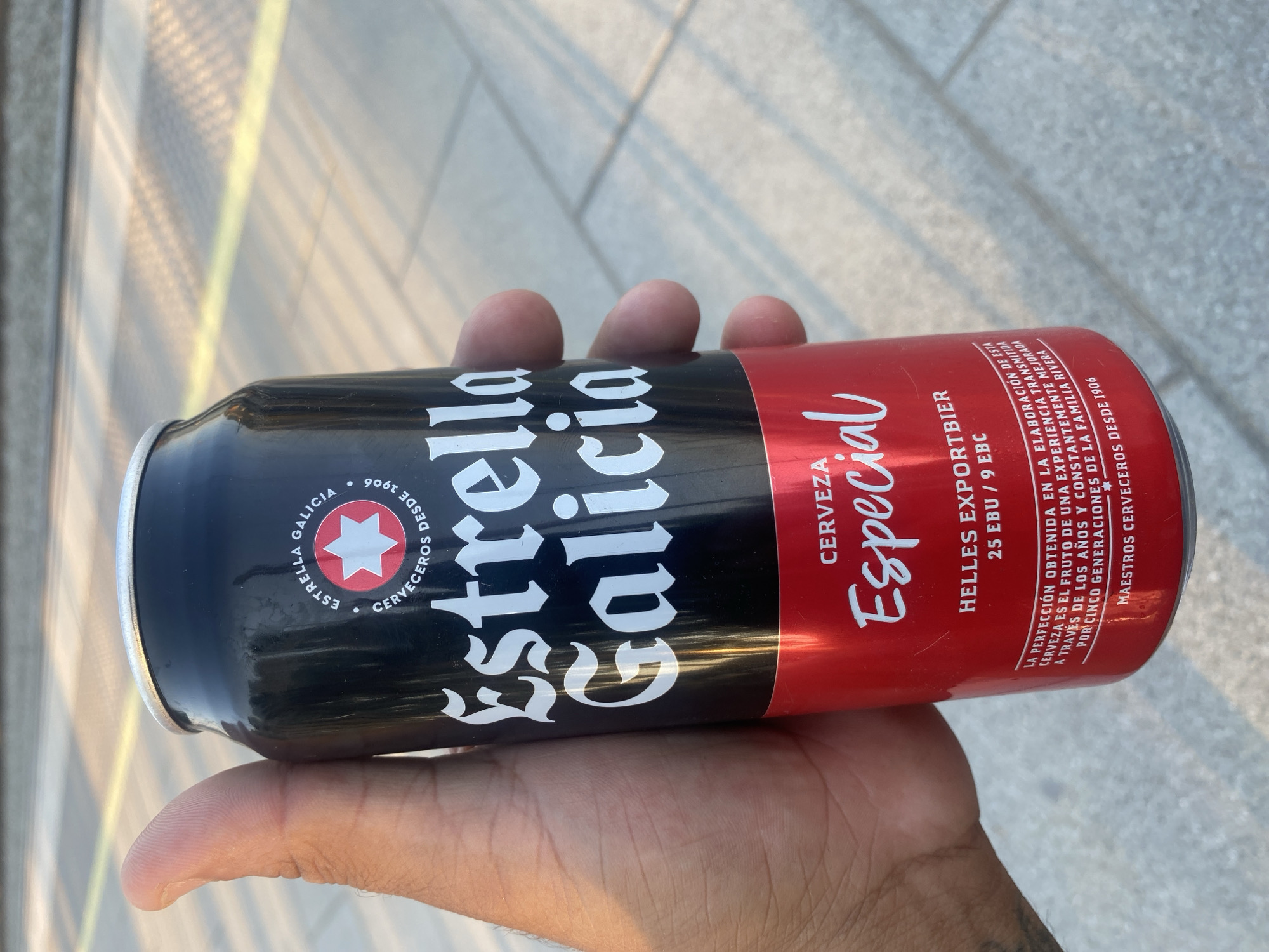 A person's hand is holding a can of Estrella Galicia beer. The can is predominantly black and red, with white lettering. The dominant text on the can reads "Estrella Galicia" in large, stylized font, with "Especial" written in a script font below it. There is also text indicating "HELLES EXPORTBIER" and nutritional information. A circular logo featuring a star within a red circle is visible on the left side of the can, along with the text "ESTRELLA GALICIA" and "CERVECEROS DESDE 1906." The can appears to be slightly wet, suggesting it might be cold.
The background is blurred, but appears to be an outdoor setting with paved ground and some vertical lines that could be part of a structure or railing. Sunlight is casting shadows, indicating it is daytime. The overall impression is casual, possibly a moment of enjoying a drink outdoors. The text on the can suggests the beer is a Spanish brand, with a long history. The location context of "la Llagosta, Spain" further grounds the scene as being in Spain. No specific event is evident, but the image captures a simple, everyday moment.