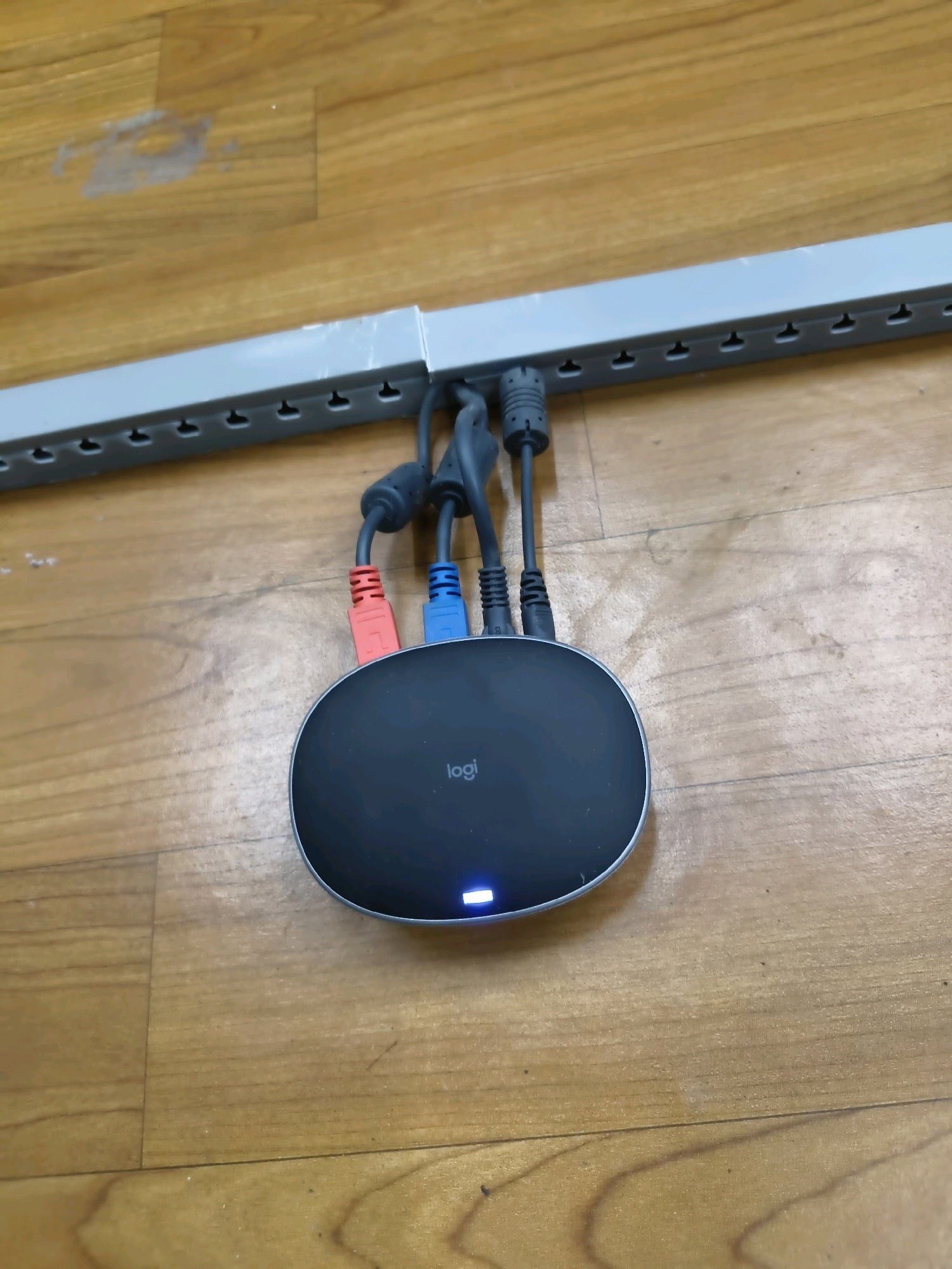 A black, oval-shaped Logitech device with a silver trim is positioned on a light brown wooden floor. Three cables, red, blue, and black, are plugged into the device. The device has a blue LED light illuminated on its front. Above the device, a gray, plastic rail with a series of notches runs horizontally across the image. The wooden floor shows some scuff marks and a patch of what appears to be dried liquid. The scene is lit from above, casting a slight shadow from the rail onto the floor. The location is not discernible from the image.