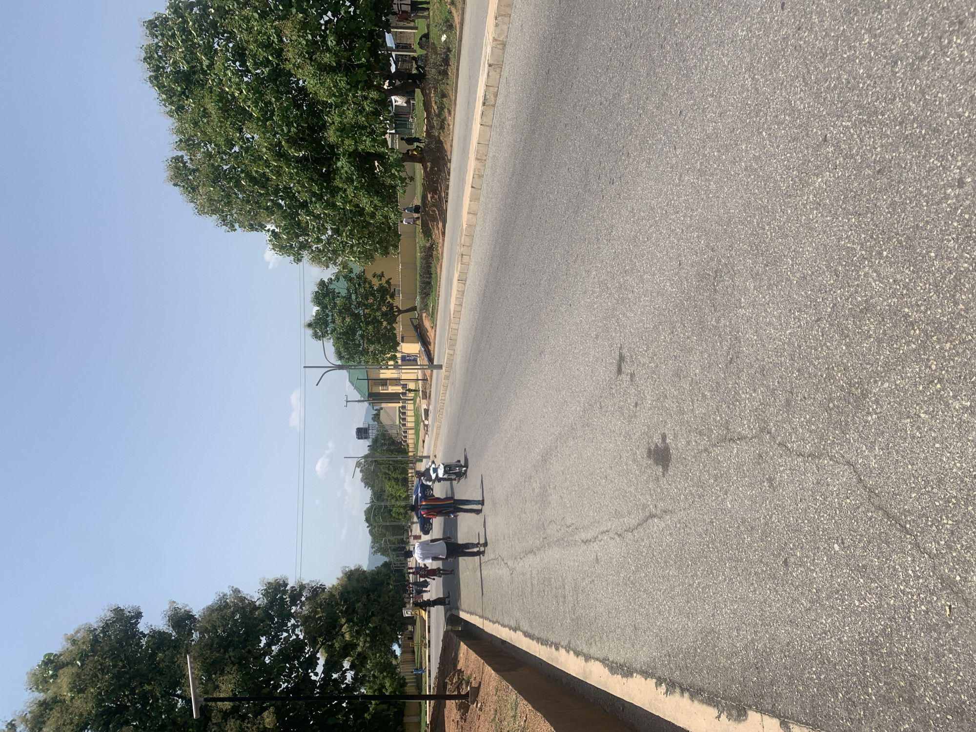 The image captures a street scene in Jalingo, Nigeria, under a clear blue sky. A paved road dominates the foreground, with a slight curve to the right. On the left side of the road, a raised dirt embankment with a concrete curb runs alongside. Trees with dense green foliage line the embankment.
Several individuals are present on the road. In the middle ground, two men are walking away from the camera. One wears a white shirt and dark pants, while the other is dressed in a striped shirt and dark pants. To their right, a person is riding a white motorcycle. Further down the road, a blue car is visible, and a group of people are walking on the left side, some in traditional attire.
In the background, buildings with light-colored walls and green roofs are visible, set back from the road and partially obscured by trees. Electrical poles and wires are also present. The overall environment suggests a suburban or semi-urban setting with active pedestrian and vehicular movement.
