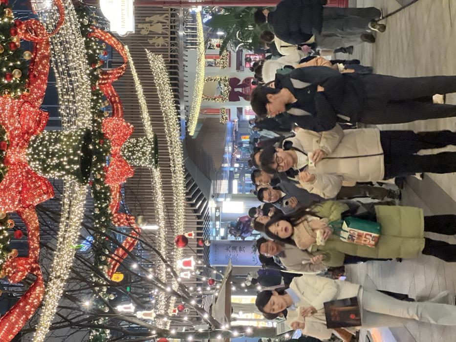 The image depicts a nighttime scene on a pedestrian street or commercial outdoor area in Liangjiang, China, heavily decorated with festive lighting. Overhead, numerous strings of warm white lights are strung in parallel lines and arches, interspersed with large illuminated red bows and bell-like structures also covered in lights. On the left, bare tree branches are adorned with white string lights, red spherical ornaments, and white bell-shaped lights. Building facades are visible on both sides of the street, with a partially visible sign on the right reading "HAPPY HIL..." above silhouettes of figures.
Approximately 20-30 individuals, predominantly East Asian, are present in the scene, moving along the thoroughfare. Most are dressed in winter attire, including puffer jackets and long coats.
In the foreground:
- A female on the far left, wearing a white jacket over a light blue top and light-colored pants, holds a small white container in her right hand and a brown paper bag in her left.
- To her right, a female wearing a light green puffer coat and a light-colored scarf holds a white container in her right hand and a green shopping bag in her left. Her mouth is slightly open, and she looks forward.
- To her right, a male wearing a light beige puffer jacket and dark pants, with glasses, holds a white container in both hands and looks down at it.
- On the far right, a male wearing a dark blue hoodie/jacket and dark pants, with glasses, stands with his arms crossed, a dark backpack visible on his back. He looks towards the right.
In the midground and background, other individuals are walking or standing. Several are observed holding and looking at smartphones. The overall action involves pedestrian movement and casual consumption of food or beverages from containers. There are no overt direct interactions between the primary foreground subjects.