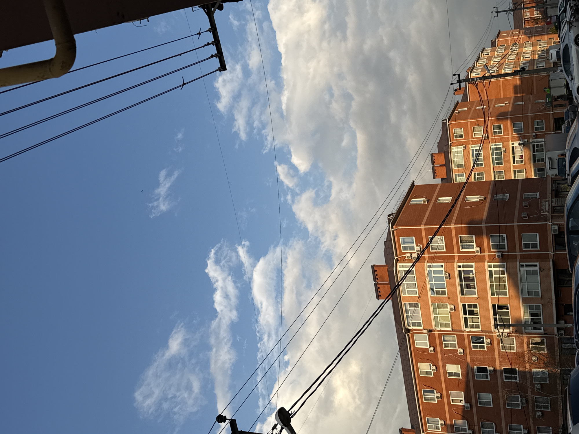 The image shows a view looking upwards at a slight angle. In the background, there are tall brick buildings with multiple windows and air conditioning units. Power lines stretch across the scene. The sky is visible, with a mix of blue sky and white clouds. A pipe runs along the top left corner. The image appears to be taken in an urban environment, in the city of Unknown, Unknown.