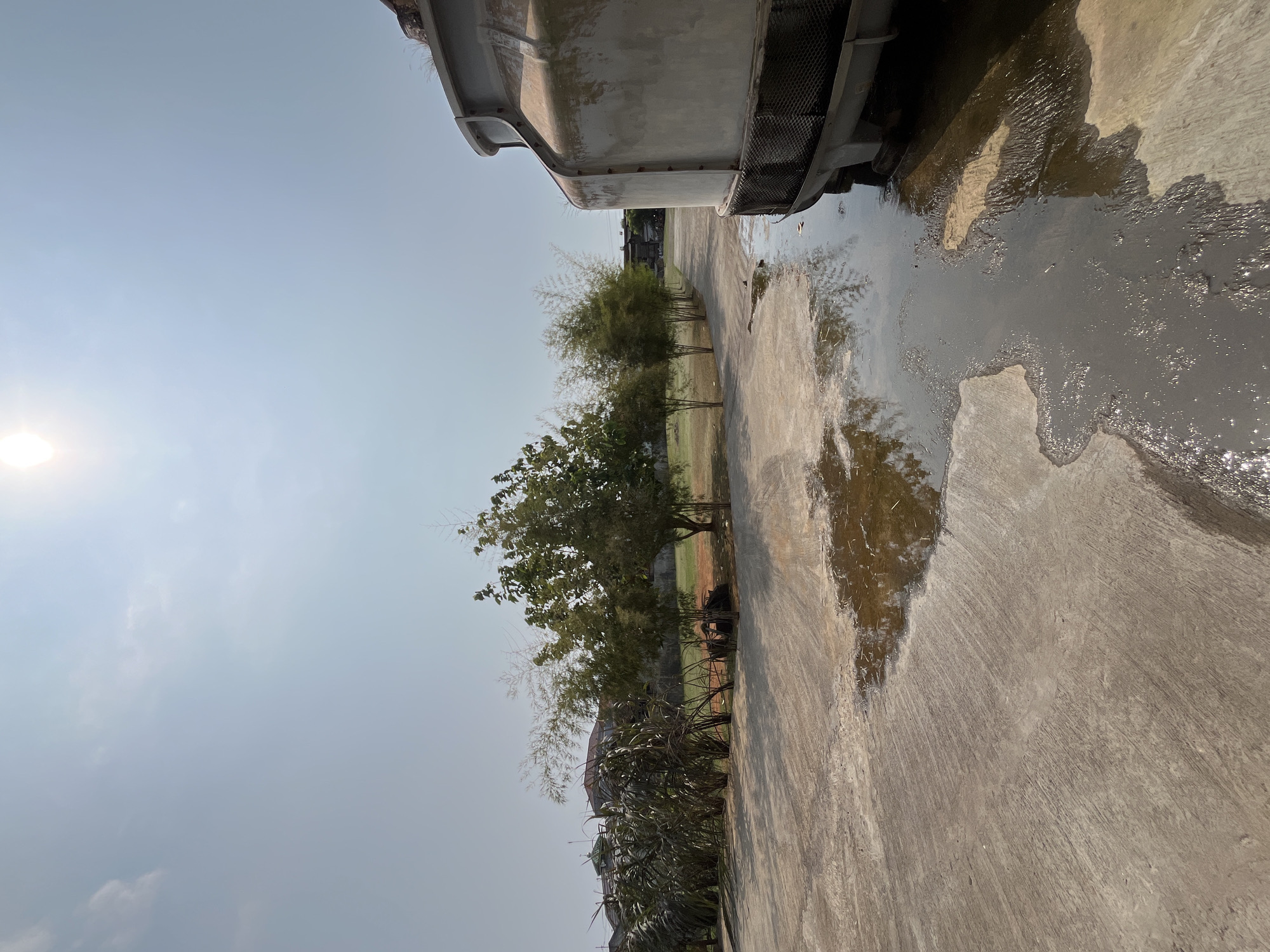 A wide-angle, low-angle outdoor shot captures a scene on a sunny day, likely in Pekalongan, Indonesia. In the foreground, a large, grey, cylindrical industrial structure, possibly a water cooling tower, dominates the right side of the frame. Its textured surface suggests wear and tear, and a black metal grille is visible at its base. A large puddle of water, reflecting the sky and surrounding vegetation, covers a significant portion of the concrete ground in the foreground and midground. The concrete itself appears weathered, with visible marks and a rough texture.
To the left of the industrial structure, a pathway or road stretches into the distance. Along this path, a line of trees and tall, grass-like plants creates a verdant barrier. Among these are several slender, bamboo-like trees with feathery foliage, and darker green, fuller trees. In the background, houses with reddish-brown tiled roofs are visible, interspersed with more vegetation and a fence.
The sky is a pale blue with a bright, hazy sun near the top center, indicating a clear and possibly hot day. The overall impression is one of an industrial or utility area adjacent to a residential or semi-rural setting. No people are visible in the image.