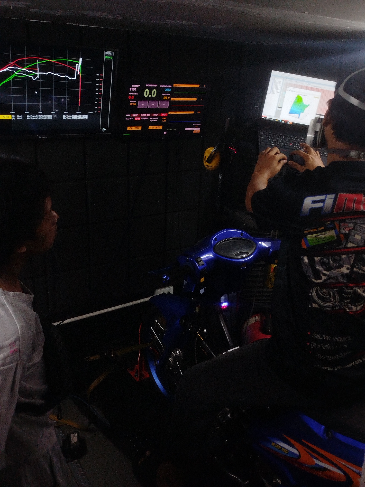 The image depicts an indoor scene, likely a workshop or garage, in Ban Kho En, Thailand, focused on motorcycle performance tuning. Two individuals are present. On the left, a person with dark, curly hair is seen from the side, wearing a light-colored shirt. They appear to be observing the equipment. On the right, a person wearing headphones and a black t-shirt with graphics is seated, operating a laptop. Their hands are visible on the keyboard.
In the foreground, a blue motorcycle is partially visible, suggesting it is the subject of the tuning process. The environment is dominated by electronic equipment. Two large monitors are displayed prominently. The left monitor shows a graph with multiple colored lines, likely representing engine performance data such as RPM, horsepower, or torque. The right monitor displays numerical data, including "TARGET 2100", "POWER HP 0.0", and "ENGINE RPM 2062". Another screen to the right shows a graphical representation, possibly a dyno graph or a map of performance parameters. Cables and various electronic components are visible around the equipment. The lighting appears to be artificial, creating a somewhat dim and focused atmosphere. The background is dark, possibly a sound-dampened wall.