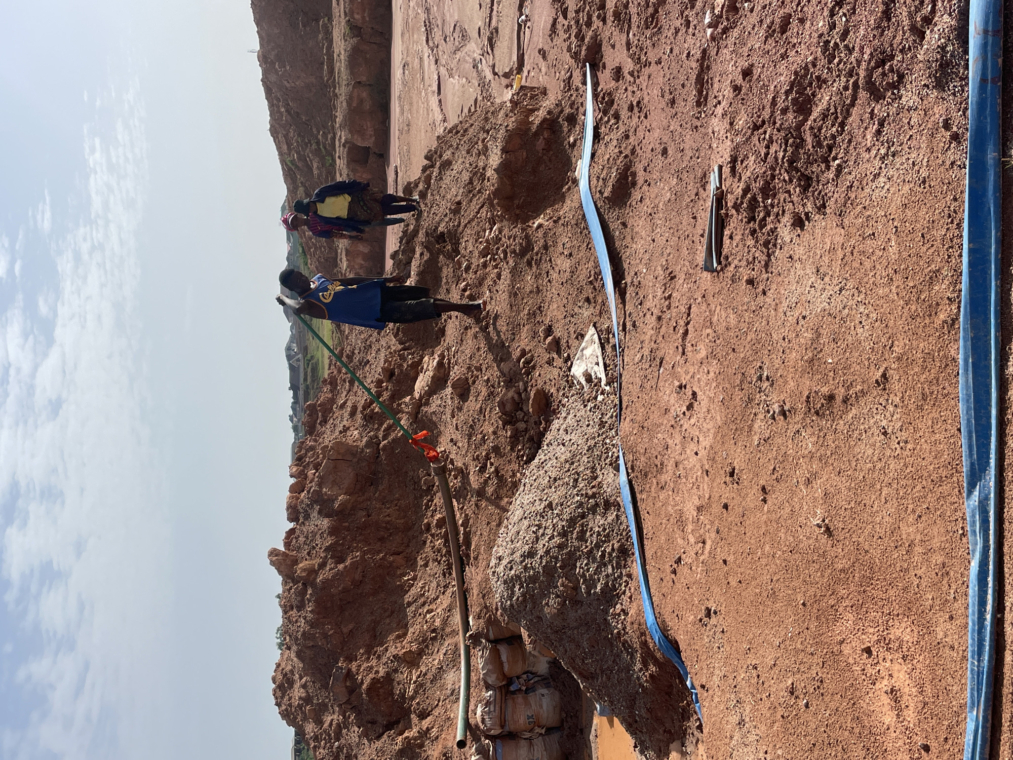 The image shows an outdoor scene likely in a rural or semi-rural area with reddish-brown soil, possibly in the city of Jos, Nigeria. There are three individuals present. One person is standing near a green pipe and hose, holding onto it, dressed in a blue shirt. Two other people stand further back on a higher part of the terrain, near what looks like a raised area or cliff.
Several objects are visible. A blue pipe or hose runs across the ground. There is a large pile of earth or a rock formation covered in loose material. Bags are visible stacked under the large pile. Additionally, a sharp metal object is visible on the ground near the blue hose.
The terrain is uneven, with areas of loose soil and what appear to be excavated areas. The sky is visible in the background, suggesting an open environment. There is no clear indication of specific activities, but the presence of the pipe and excavation suggests digging or water access may be occurring.
