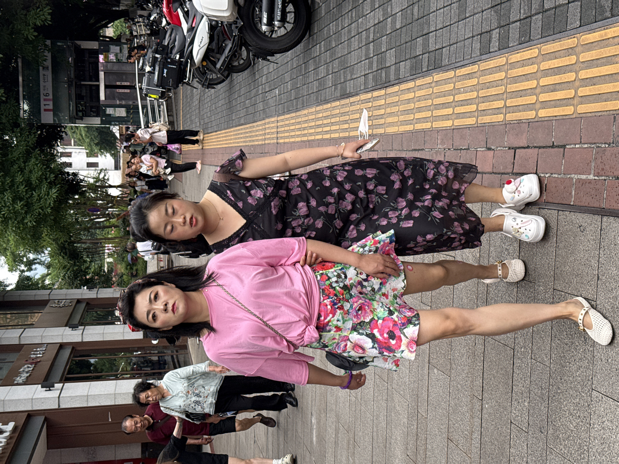 Two Asian women stand on a sidewalk with a tactile paving strip along the edge. One woman wears a black floral dress and white Crocs. The other wears a pink shirt, a floral skirt, and white sandals. They appear to be posing for a picture. In the background are pedestrians, buildings, and parked motorcycles. The location is urban. Based on the context, this is likely in the Yuzhong District, China.