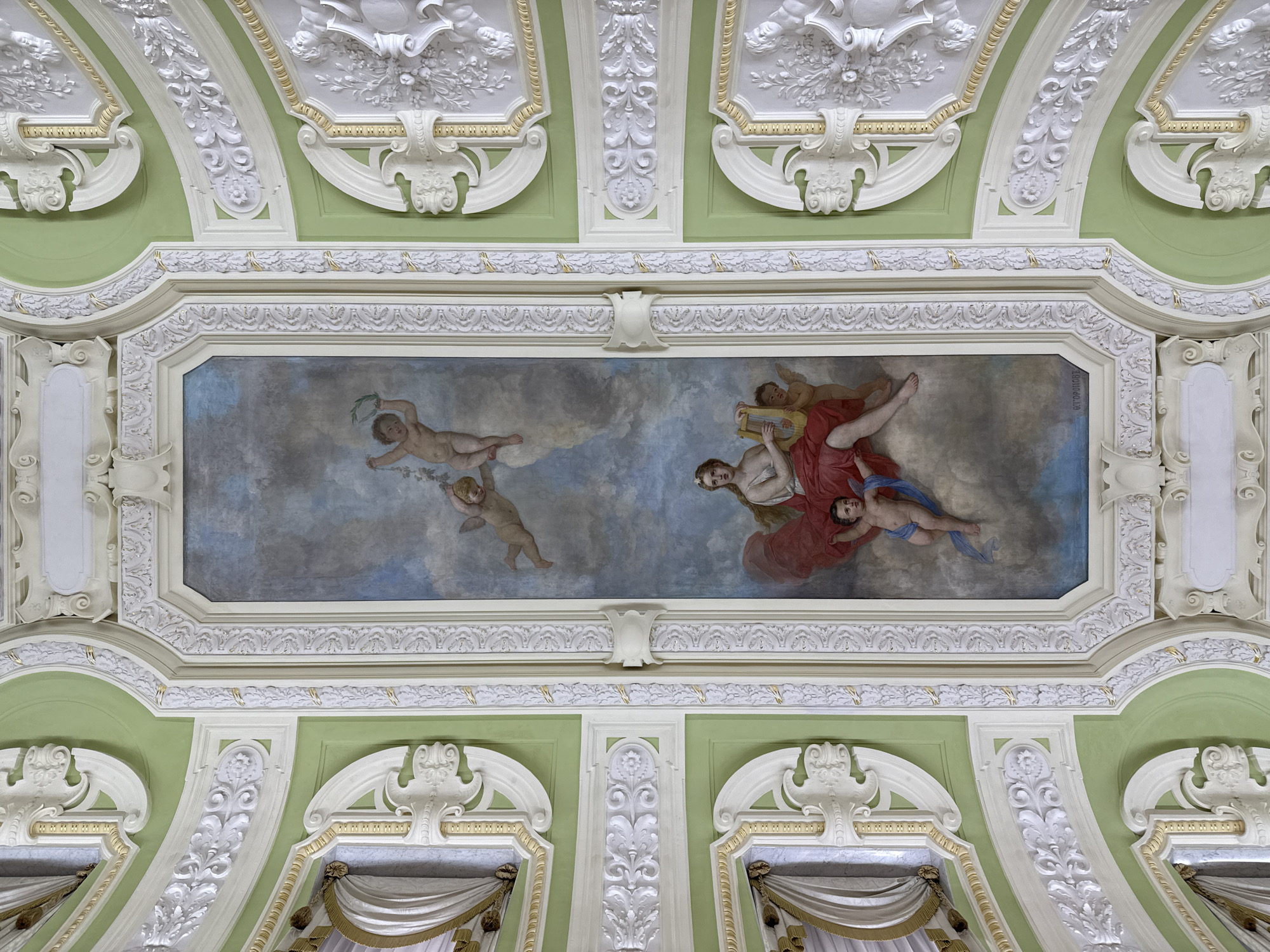 The image shows a frescoed ceiling in the Nizhny Novgorod Kremlin, Russia. The fresco depicts a scene with mythological figures. In the center, a woman with long blonde hair, dressed in a red robe, plays a lyre. She is surrounded by cherubs. One cherub is flying above her with a laurel wreath, while another is reaching out to her from below. The ceiling is decorated with intricate stucco moldings, painted in pale green and white, with gold accents. The fresco is framed by ornate white and gold borders.