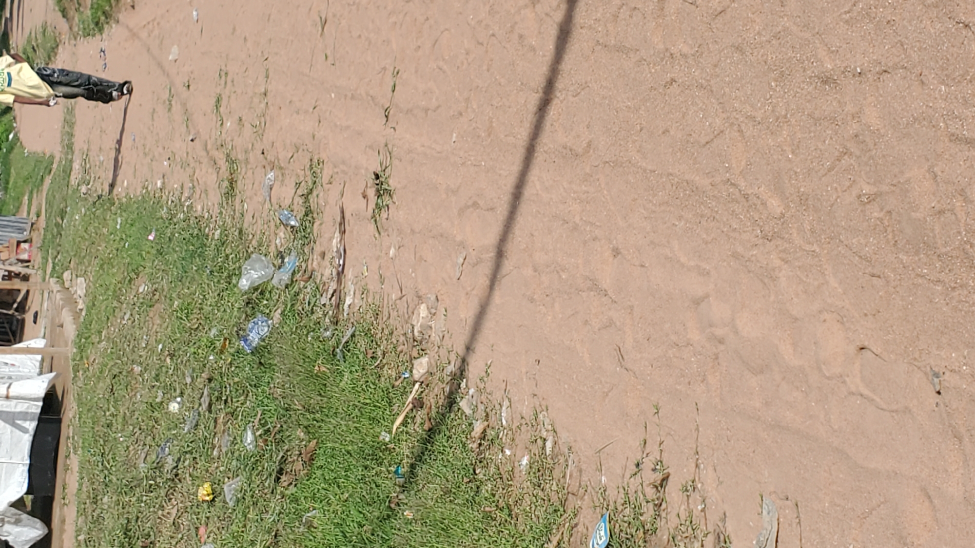 The image depicts a scene in Jalingo, Nigeria, characterized by a dirt path with sparse vegetation and scattered litter. On the right side of the path, a person, likely a male, is walking away from the camera. They are wearing a yellow t-shirt with an emblem on the front and dark, patterned trousers, along with dark shoes. The path itself is composed of reddish-brown sand, with tire tracks and footprints visible. To the left of the path, there is a patch of green grass, also littered with plastic bottles and other debris. In the background, there are some structures that appear to be makeshift shelters or stalls, covered with white tarpaulins and corrugated metal sheeting, suggesting a semi-urban or rural setting within the city. The overall impression is one of a well-trafficked but unkempt area.