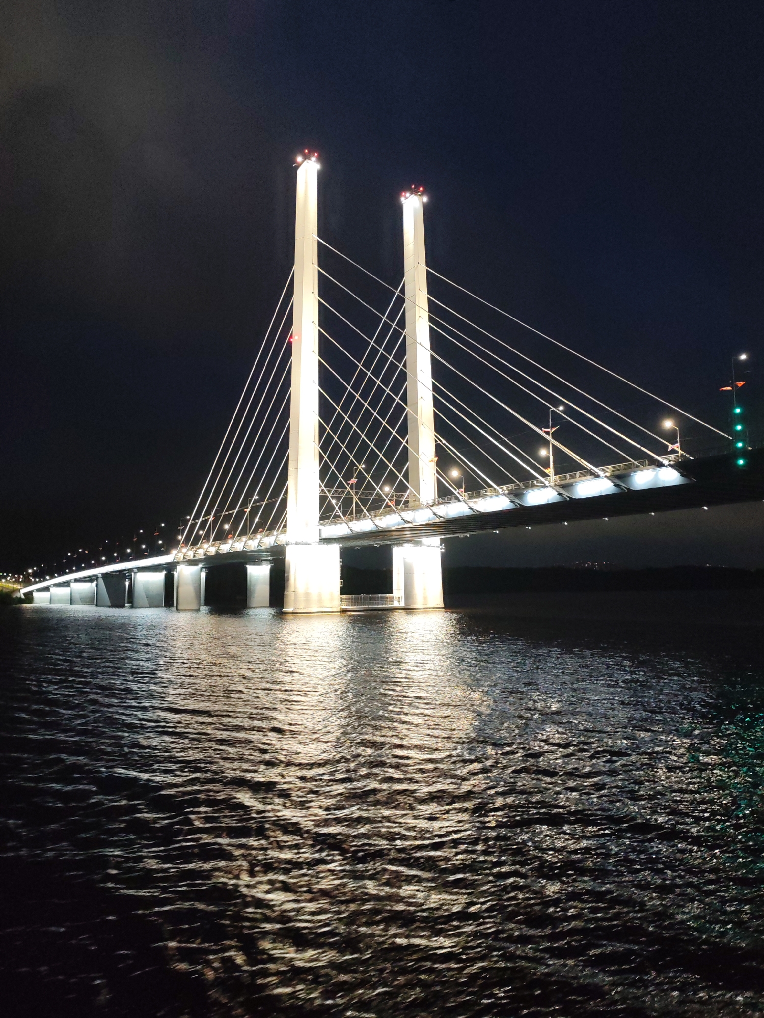 At night, a cable-stayed bridge is illuminated with white lights and red warning lights on the towers. The cables stretch from the towers to the bridge deck. The bridge deck is also illuminated with white lights. The water below the bridge reflects the lights, creating a shimmering effect. The environment is dark and the sky is black.