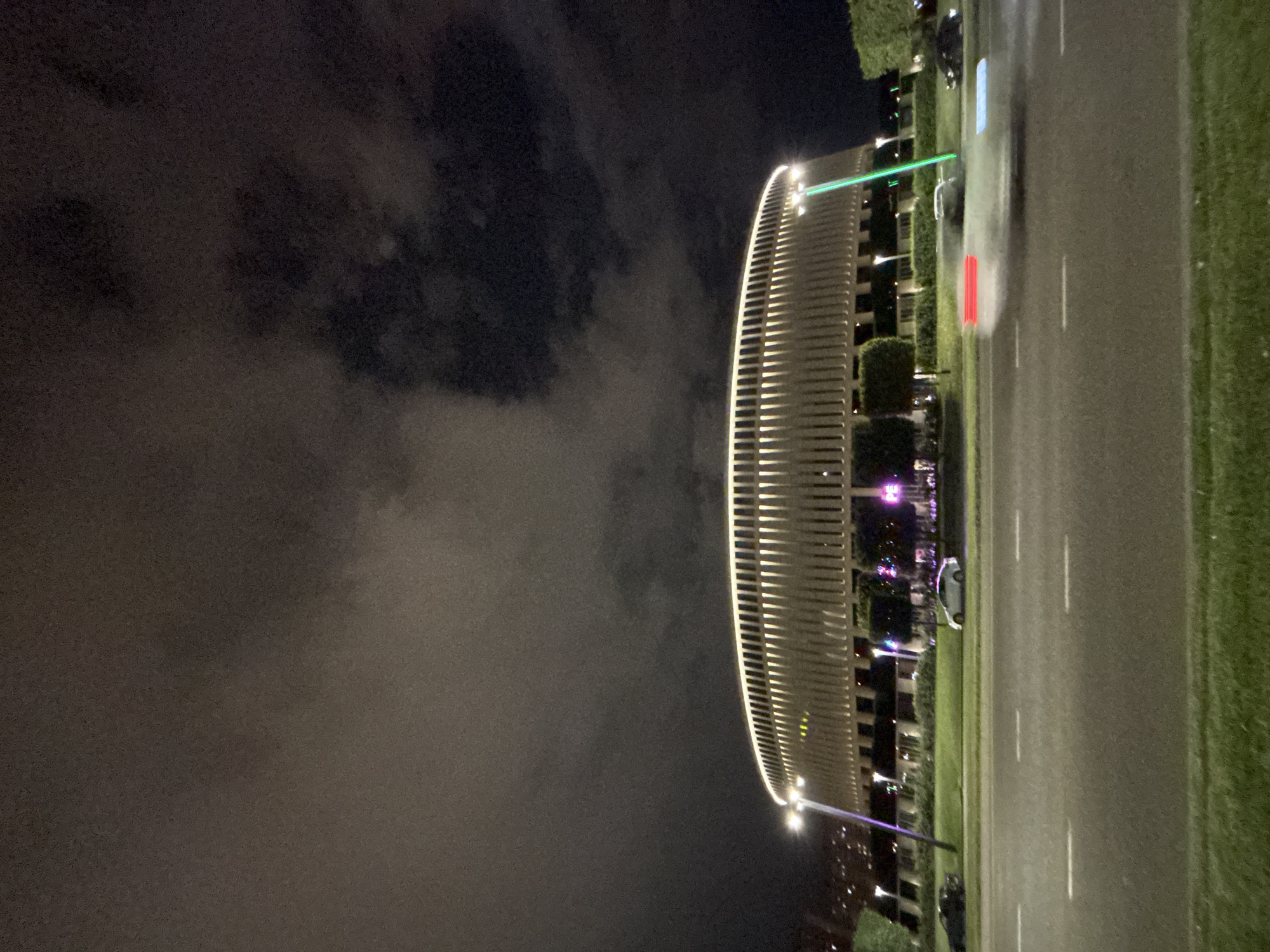 The image, taken at night in Krasnodar, Russia, shows a modern, curved building illuminated with warm lighting. The building appears to be an office or residential complex, with a sleek, contemporary design. A bright green laser beam is visible, projecting from the building across the scene. Adjacent to the building is a road with vehicle headlights and taillights creating streaks of light, suggesting movement. There is a grassy area bordering the road. The sky is dark and cloudy, indicating overcast weather. The overall mood is somewhat futuristic and dynamic due to the lighting and laser beam. There is no visible text in the image.