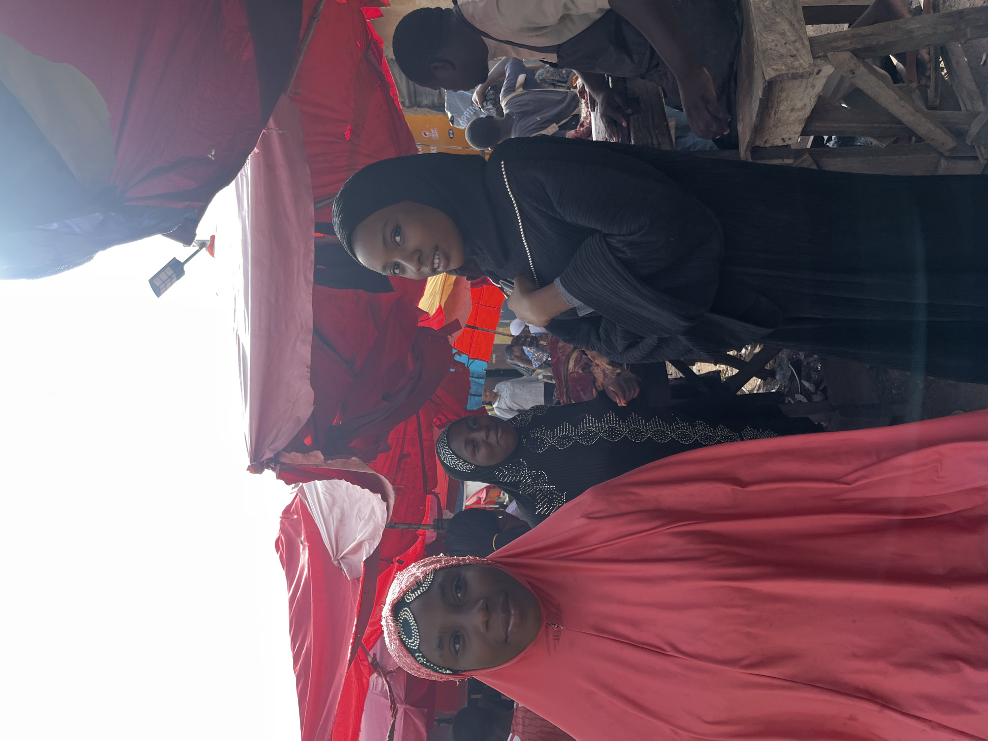 The image depicts an outdoor market scene in Rafin Par, Nigeria, under a bright sky and partially covered by red and white awnings. In the foreground, two young women are visible. The woman on the left wears a bright pink hijab and a matching garment, her face turned towards the camera. She is adorned with a beaded headdress. To her right, another young woman is dressed in a black abaya and hijab, also adorned with beads. She is looking slightly away from the camera. Behind them, other individuals are present in the market, including a man wearing a light-colored shirt and a dark apron, who appears to be working at a stall with what looks like meat. The market environment is suggested by the presence of stalls, awnings, and the general hustle and bustle.
