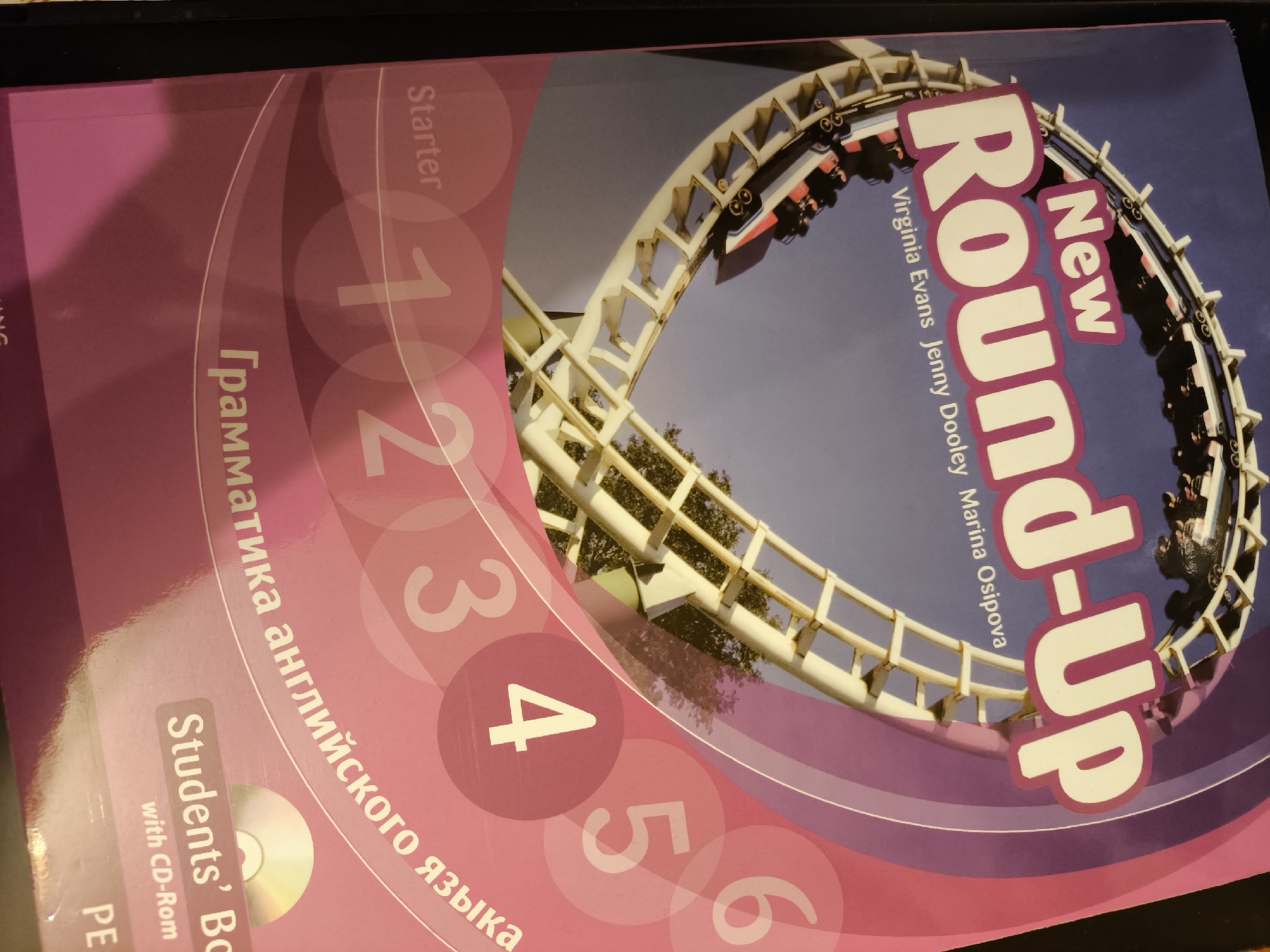 The image shows a "New Round-Up" English language textbook, specifically the "Starter" level. The book cover features a pink color scheme with stylized numbers "1" through "6" overlaid on the left side. The title "New Round-Up" is displayed prominently, along with the authors' names: Virginia Evans, Jenny Dooley, and Marina Osipova. A photo of a roller coaster track is incorporated into the design, suggesting the theme of the book. Below the numbers, there is a text written in Russian: "Грамматика английского языка", which translates to "English Grammar". It also says "Students' Book with CD-Rom" at the bottom left. There is no information in the image that indicates that the textbook relates to the city of Khimki, Russia.