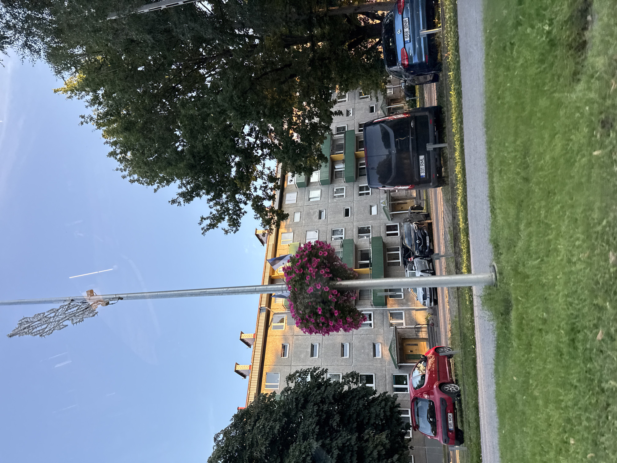 The image depicts a street scene in Tallinn, Estonia, featuring an apartment building and parked vehicles. The apartment building is a multi-story structure with a grey facade, numerous windows, and balconies, some of which are green. The building appears to be residential, with evidence of well-maintained flower boxes on a lamppost in the foreground. Several cars are parked in front of the building, including a red hatchback, a dark-colored van, and a blue sedan. The setting is an urban environment with trees and a clear blue sky overhead, suggesting it is daytime. The image has a reflective quality, indicating it may have been taken from inside a vehicle or through a window.