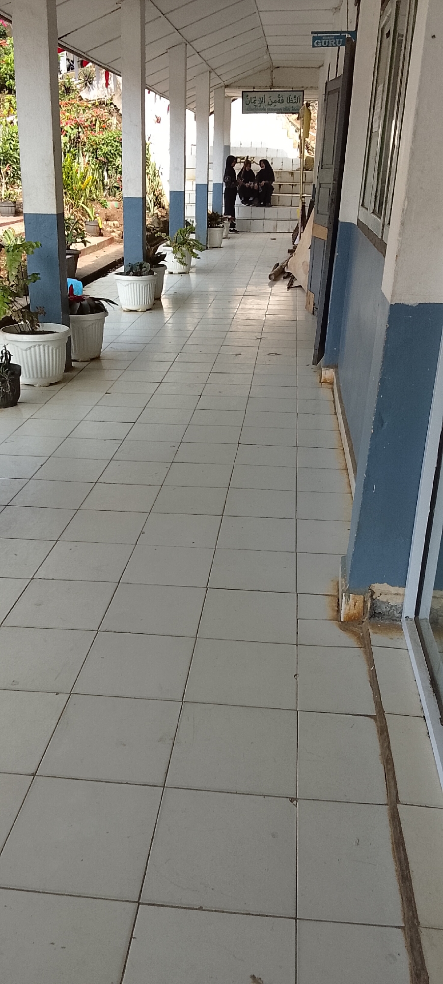The image shows a covered walkway with white tiled flooring in Jepara, Indonesia. Along the left side of the walkway, several white planters contain green plants. On the right side, a blue and white wall runs along the length of the walkway. Towards the back of the walkway, a set of stairs leads up to a doorway. Three individuals, two women wearing hijabs and one person in a black garment, are seated on the stairs. A sign with Arabic text is visible above the doorway. Another sign with the word "GURU" is mounted on the ceiling near the doorway. The walkway is illuminated by natural light filtering in from the open side.