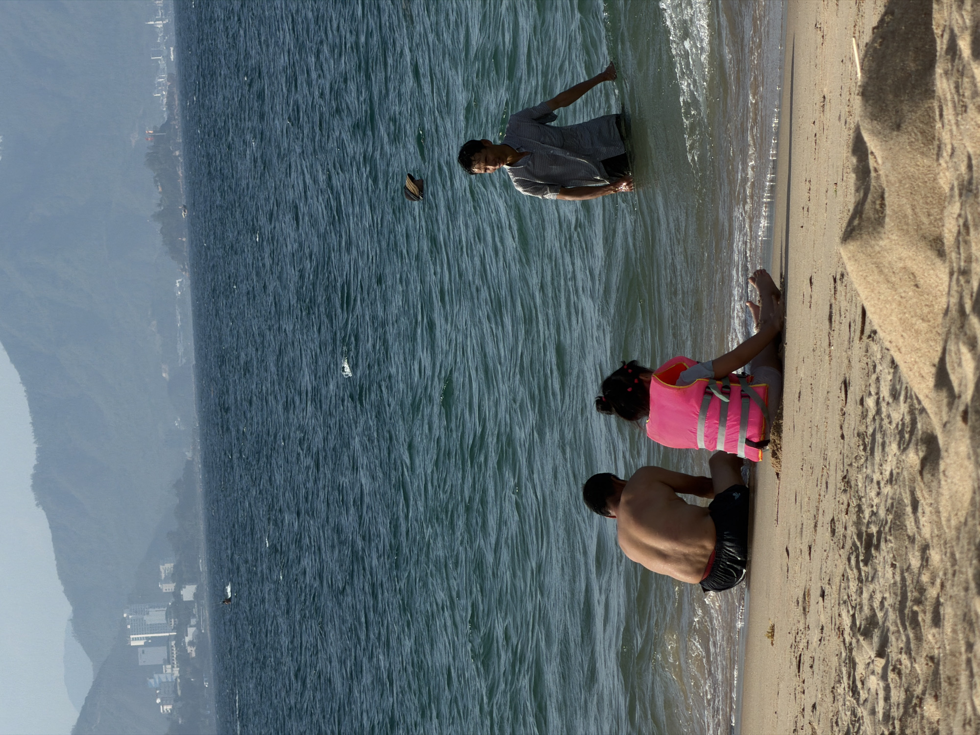 The image depicts a daytime coastal scene at Nha Trang Ward, Vietnam. In the foreground, a sandy beach occupies the lower portion of the frame, characterized by visible textures and a small, conical sand pile near the bottom center. Three individuals are positioned at the water's edge. An adult male is seated on the wet sand on the left, facing away from the viewer, wearing black swim trunks with a red waistband. To his right, a child is also seated on the sand, facing away, wearing a bright pink life vest with grey reflective stripes over a light-colored shirt and shorts. Further to the right, an adult male stands in the ocean, submerged approximately to his waist, wearing a light-colored striped short-sleeved collared shirt and dark shorts, looking towards the right side of the frame. A dark, wide-brimmed hat floats on the water's surface behind the standing male. The ocean displays small waves breaking near the shore and extends into the distance with a deep blue-green hue. In the background, a coastline unfolds, backed by hazy mountains. On the left, a cluster of white multi-story buildings belonging to a city are visible against the mountain foothills. On the right, a distinct, tree-covered rocky island or outcrop extends into the sea, featuring visible flags, with additional urban structures discernible along the distant coastline. On the far-left horizon of the ocean, a person on a personal watercraft, possibly kite-surfing, is visible creating a wake.