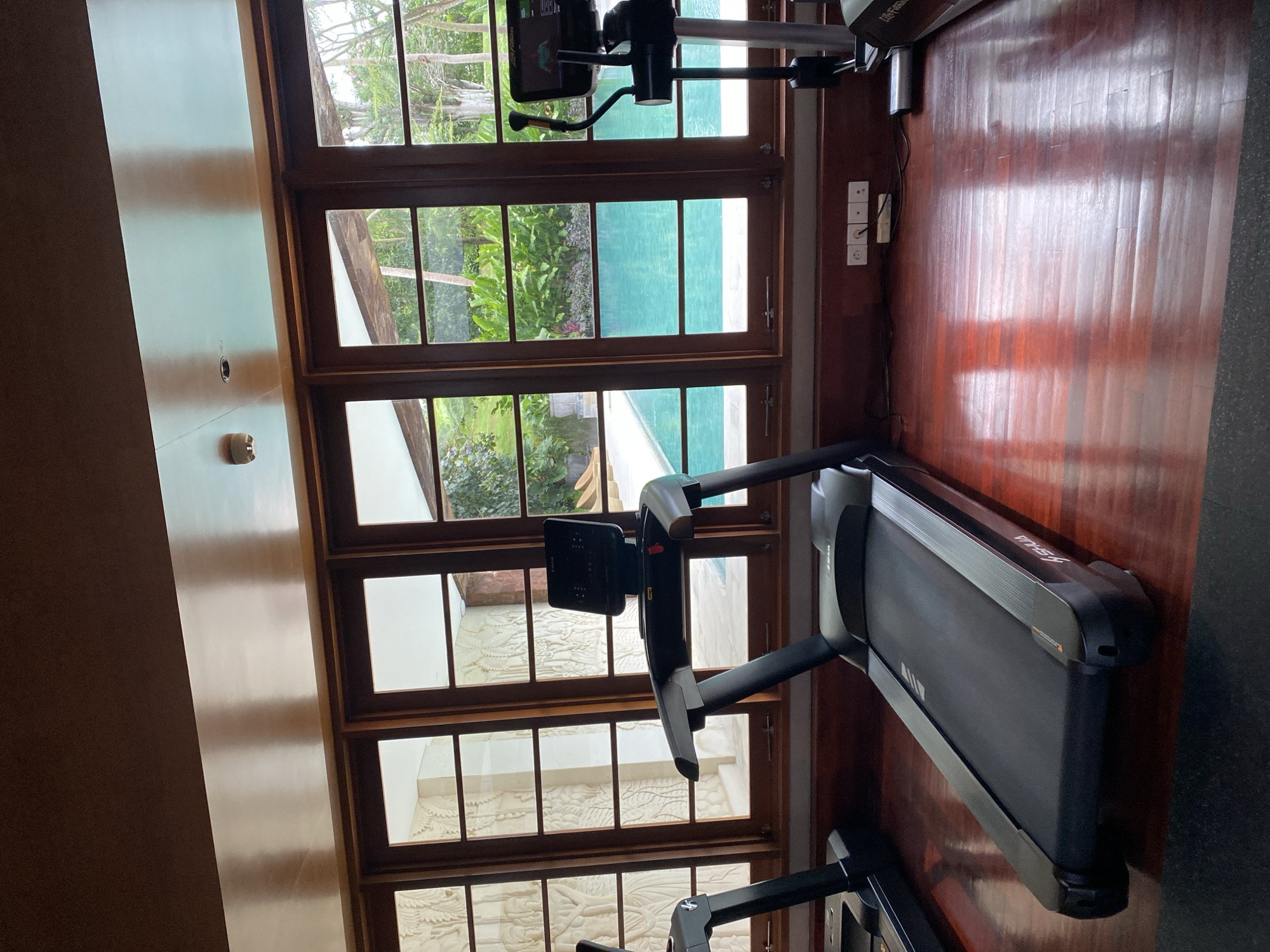 A gym in Kuta Selatan, Indonesia features two treadmills and an elliptical machine arranged on a polished wooden floor. Large windows behind the equipment offer a view of a swimming pool surrounded by lush greenery. The interior is illuminated by natural light filtering through the windows, and the ceiling has a reflective finish. The walls are adorned with textured panels. Electrical outlets and cables are visible along the base of the window frames. There are no people present in the gym.