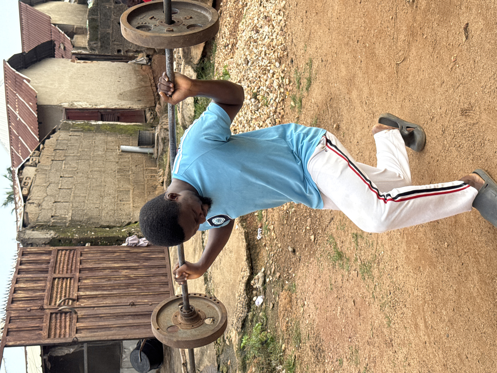 A young Black man is performing a barbell lunge exercise in an outdoor setting in Akwanga, Nigeria. He is wearing a light blue t-shirt with a white and black logo on the sleeve and white pants with red and black stripes down the sides. He has on dark grey slip-on shoes. He is gripping a barbell with both hands, which is resting on his upper back and shoulders. The barbell has two large, circular weight plates attached to each end. The ground is a mix of dirt and small stones. In the background, there is a large, rustic metal gate with vertical planks and a rounded handle. Behind the gate, there are several buildings with cinder block walls and corrugated metal roofs. Some of the buildings appear to be unfinished. There is also some sparse vegetation visible. The overall impression is of a humble, outdoor training environment in a residential area.