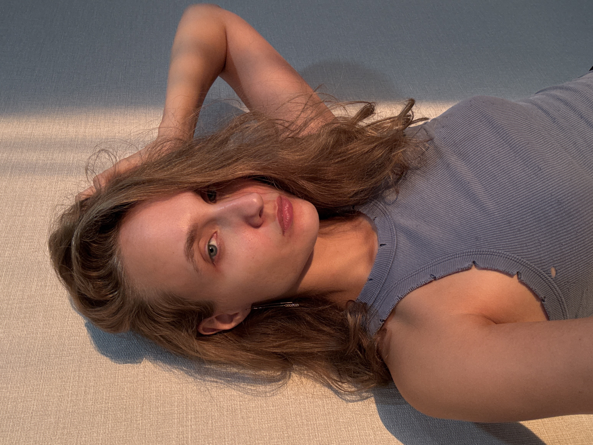 In a close-up, indoor shot, a young woman with long, light brown hair is positioned against a textured wall. She is looking towards the camera with a neutral expression, her right hand touching her hair near her temple. Her left arm is raised behind her head, and she is wearing a light blue ribbed tank top with frayed edges. The lighting suggests the presence of a window to the left, casting a warm, golden light and a distinct shadow on the wall behind her. The scene appears to be a domestic setting, possibly a home in the city of Podshivalovo, Russia.