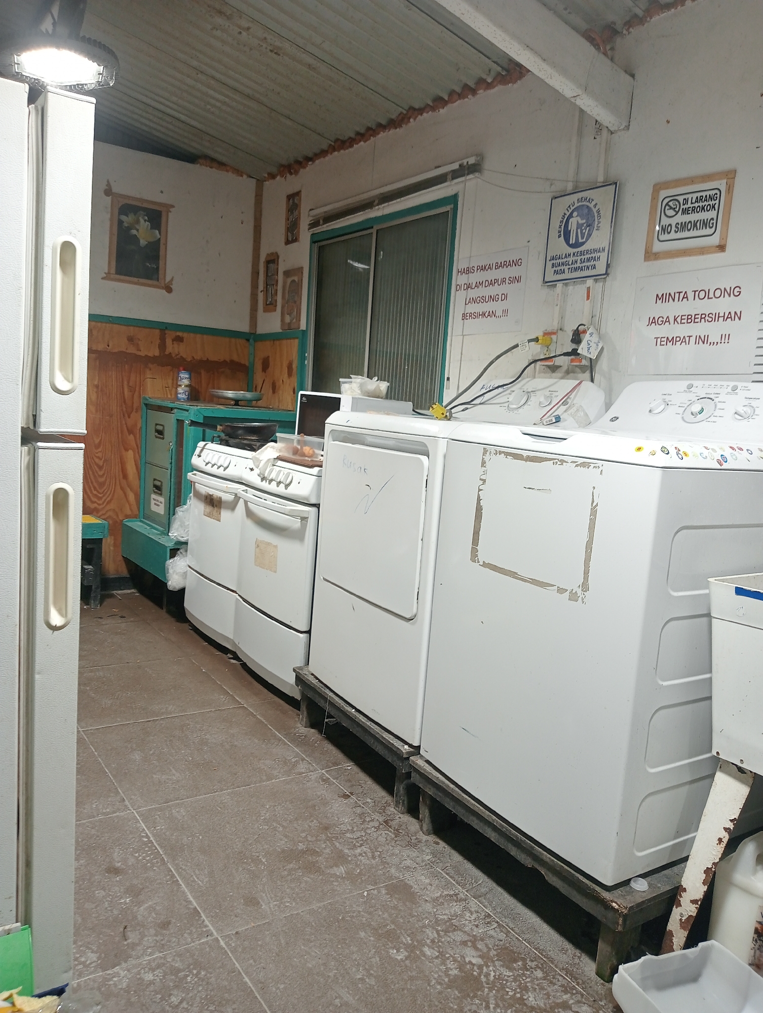 The image shows a cluttered, dimly lit room that appears to be a laundry or utility area within a dwelling. The room has a corrugated metal roof and concrete tiled floors. Appliances such as a large white refrigerator, a white gas stove, and two white washing machines dominate the space. Various signs are posted on the wall, including "NO SMOKING," a sign about keeping the area clean, and another with text in Indonesian that appears to be a reminder to clean up after use. There are also framed pictures on the wall. Wooden paneling is visible in some areas. Objects are stacked and scattered around, including plastic bags and containers. The overall impression is one of a functional but somewhat disorganized space.